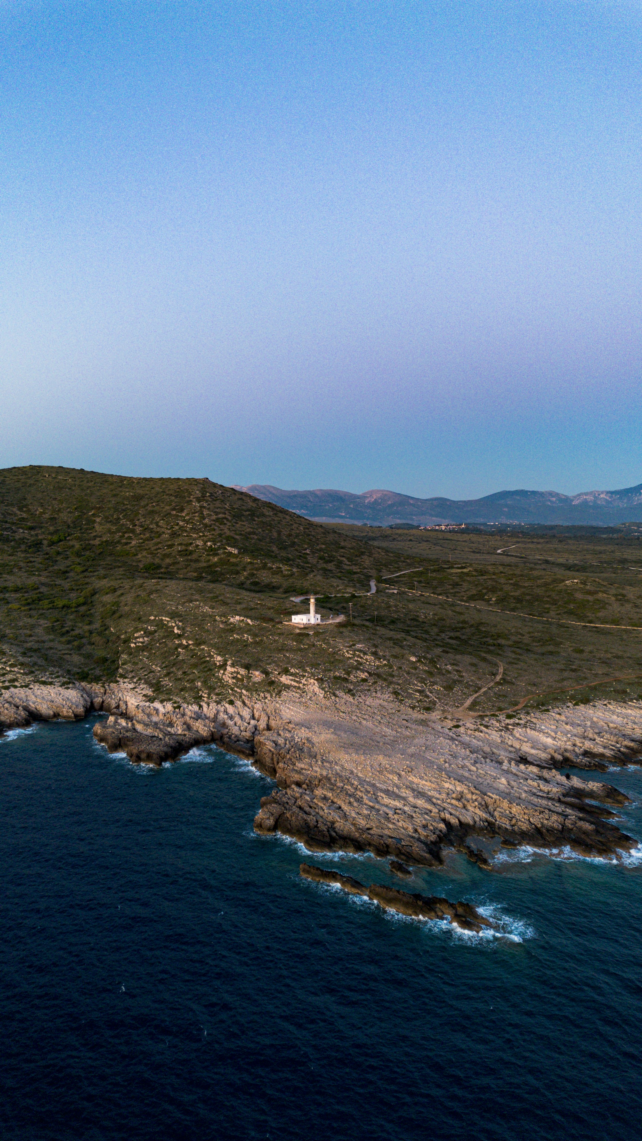 Lighthouse on rocky coastline at dusk with deep blue ocean.