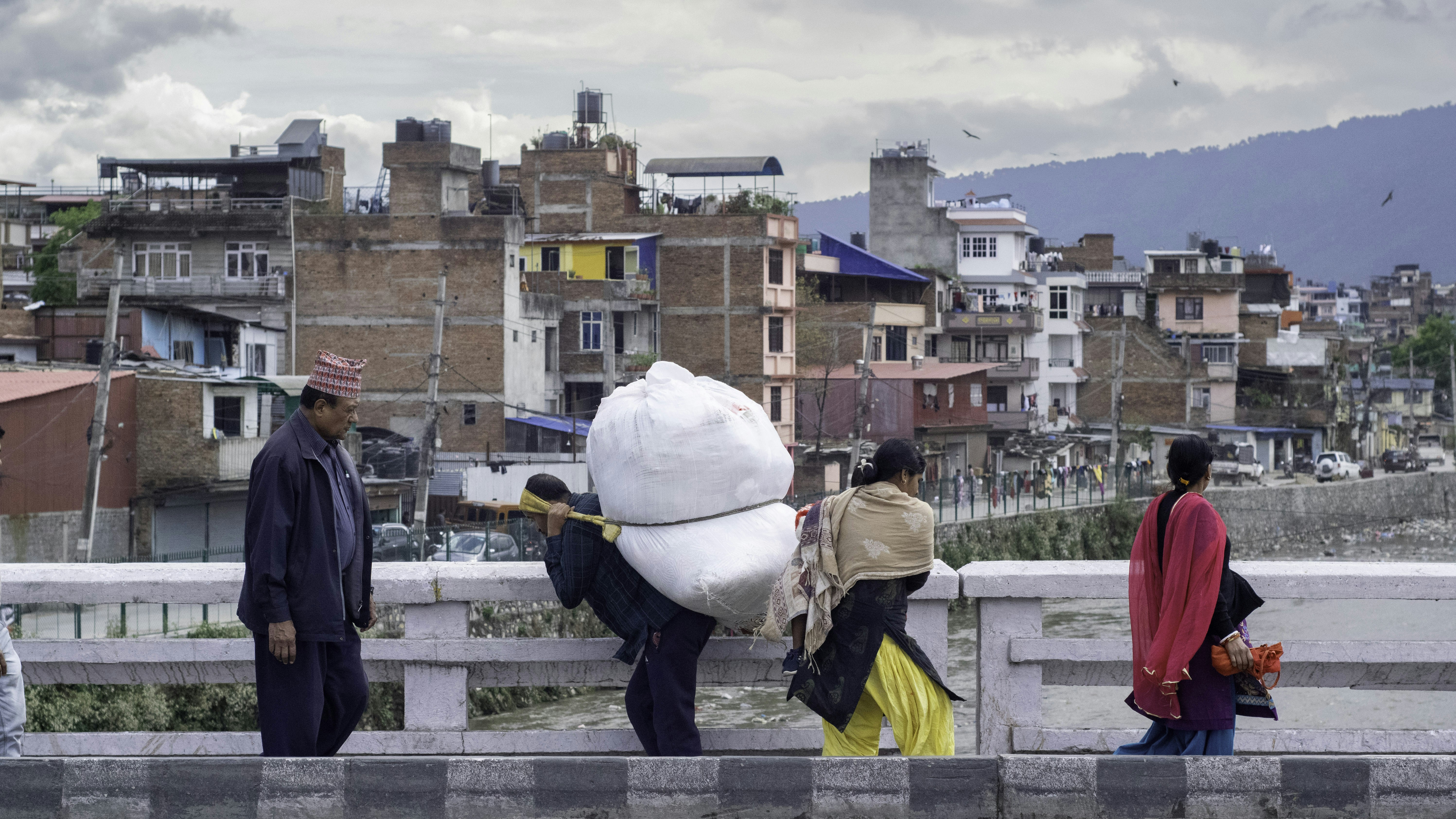 People carrying heavy loads on a bridge with buildings behind. (Photo by Dhirendra Bhatta on Unsplash)