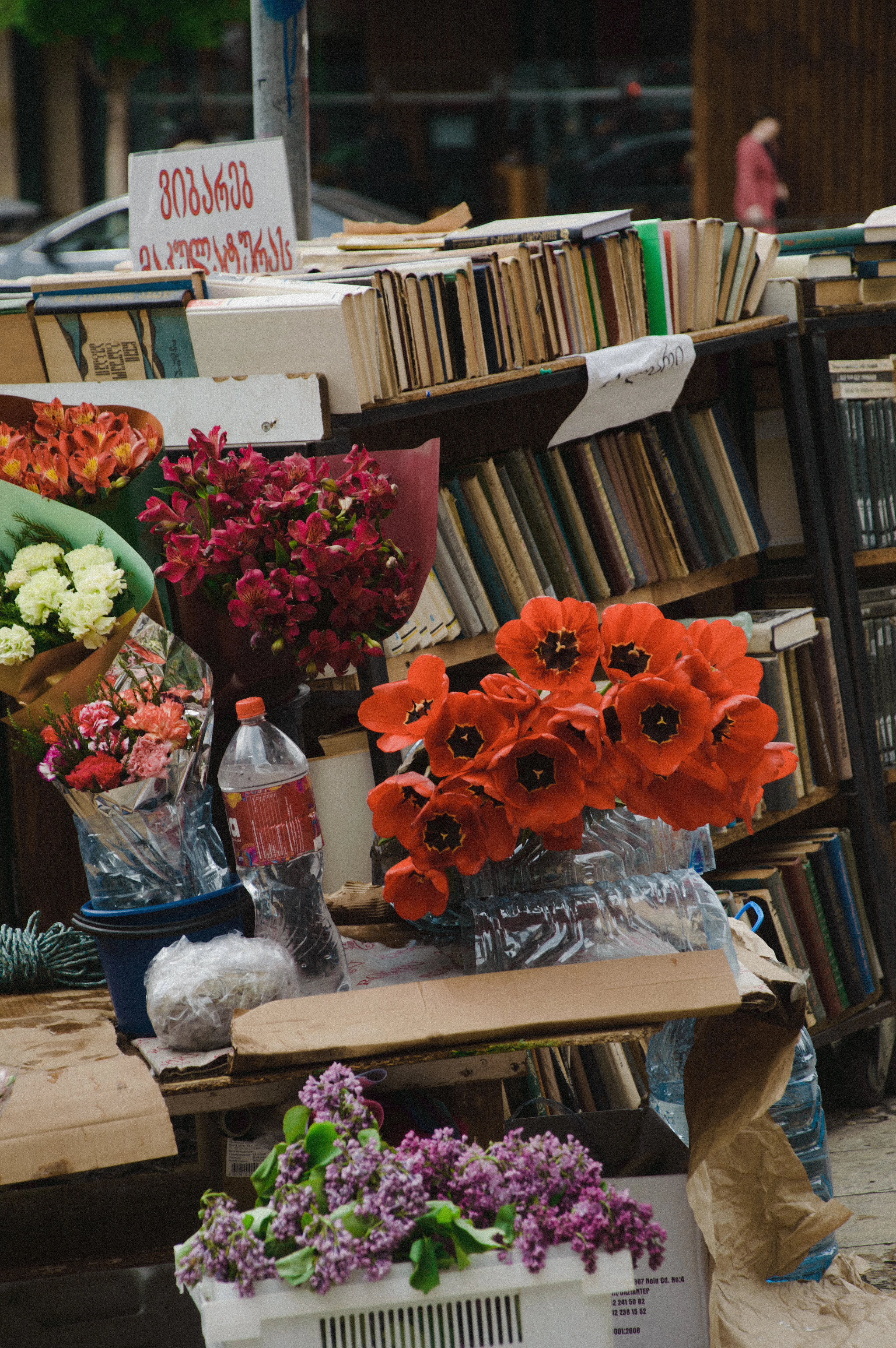 A second-hand book stall on a Tbilisi street, worn hardback spines stacked along tiered shelves with a handwritten sign in Georgian above, and buckets of cut flowers in front — scarlet tulips, pink alstroemeria, white carnations and lilac
