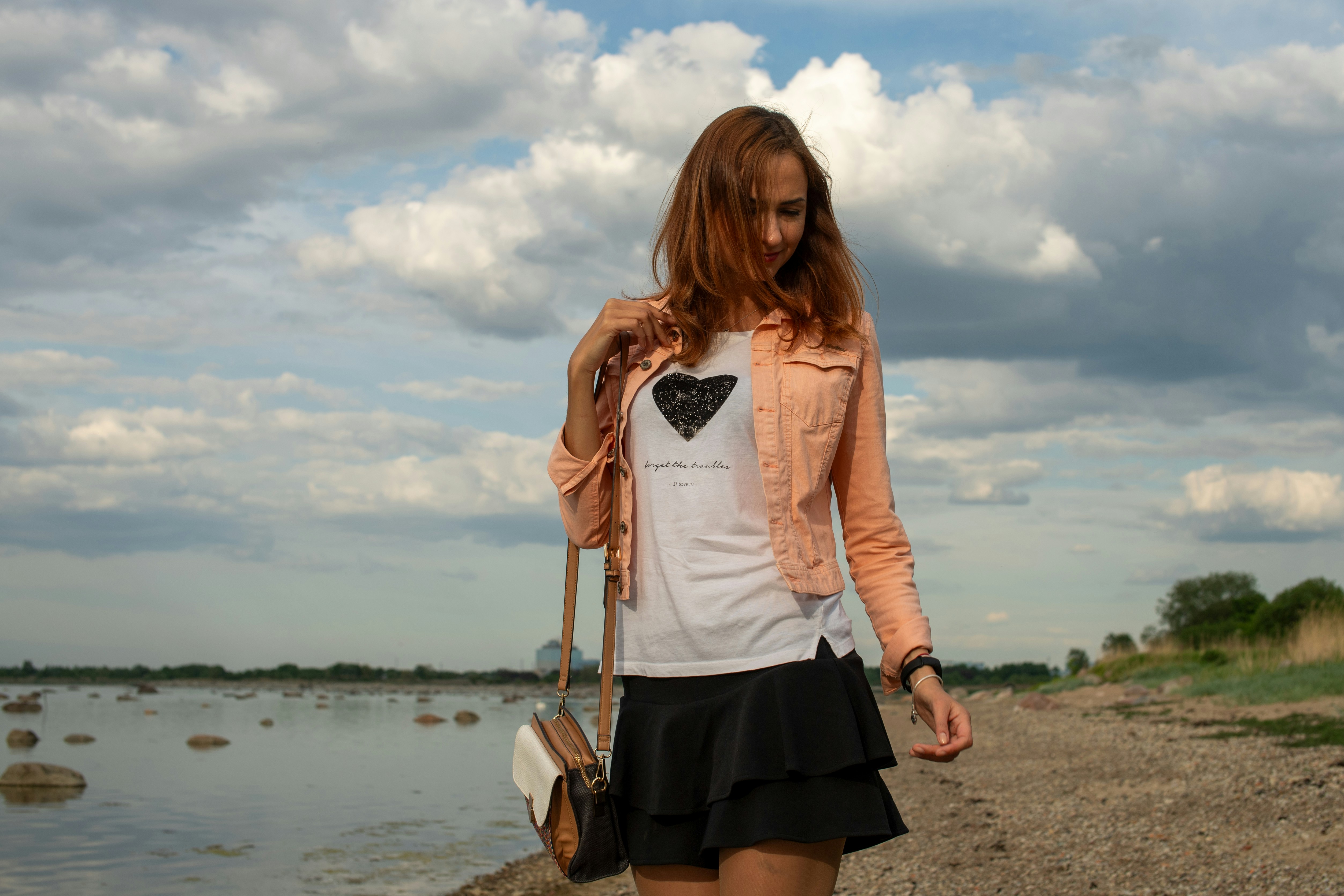 "A young woman with her hair blowing in the wind stands on the rocky shore of Kopli, Tallinn, looking out at the Baltic Sea under a dramatic cloudy sky."