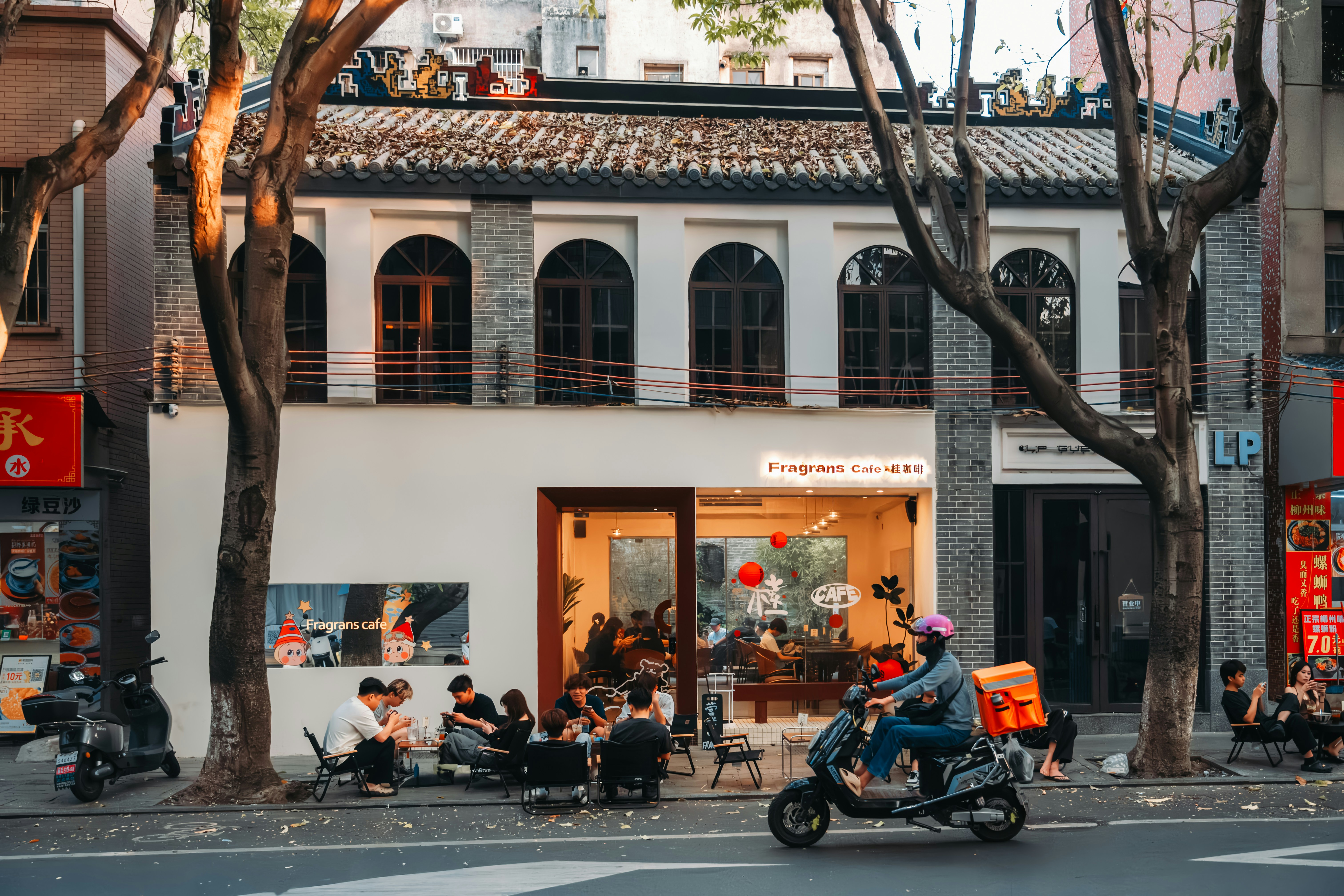 People gather outside a cafe on a tree-lined street.
