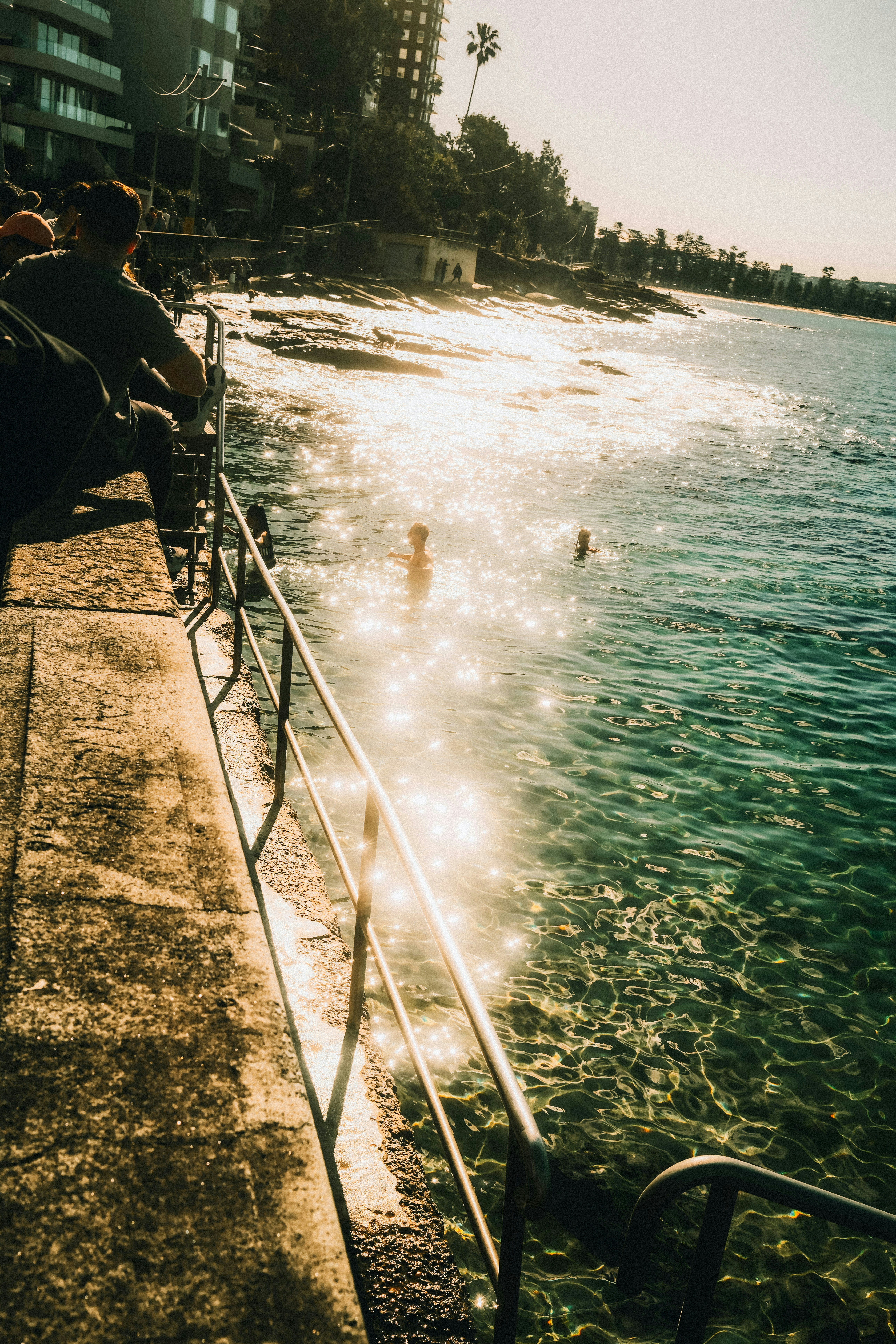 People swim in sunlit ocean water near a concrete walkway.