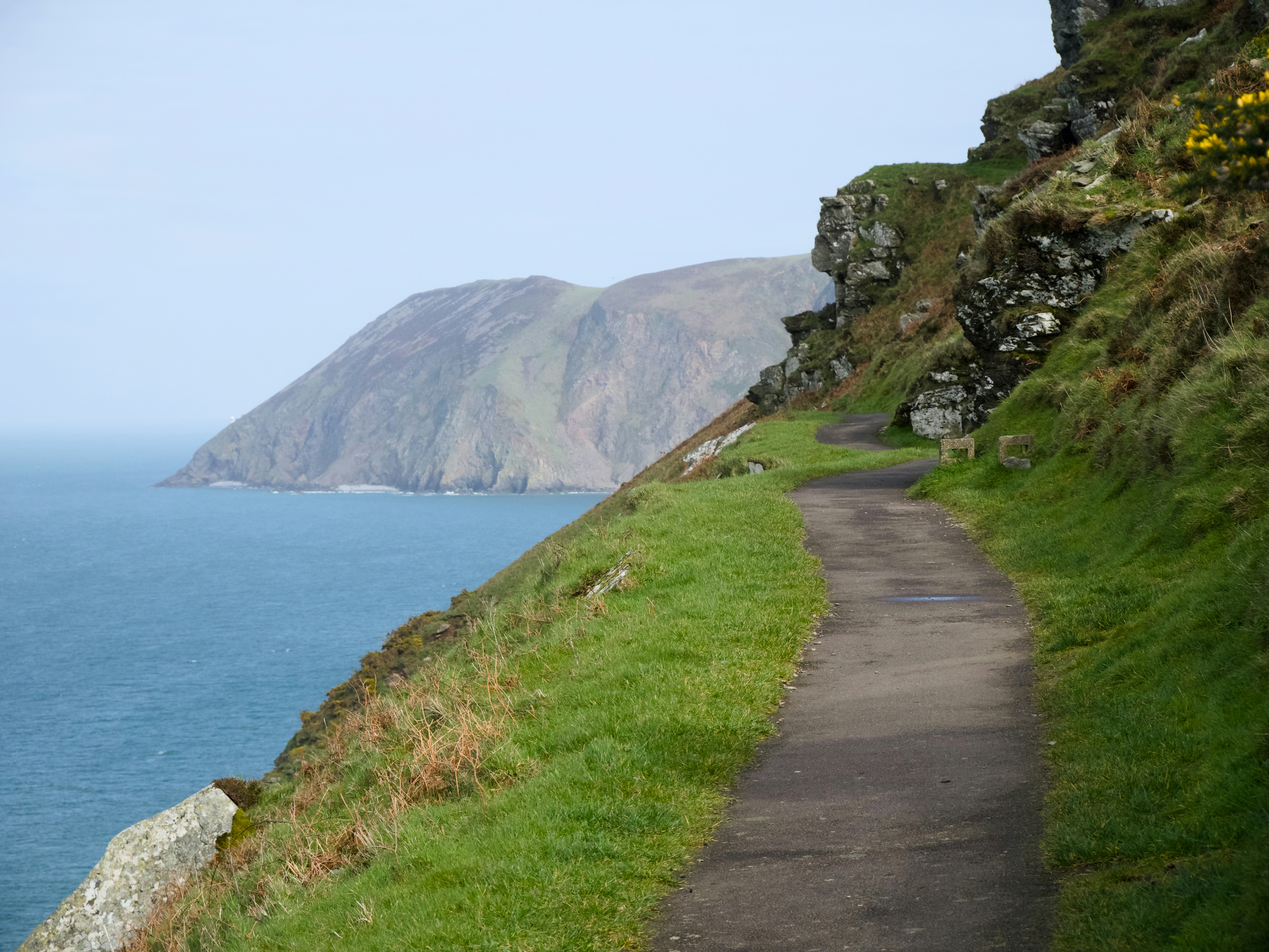 Coastal path winds alongside a grassy cliff overlooking the sea.