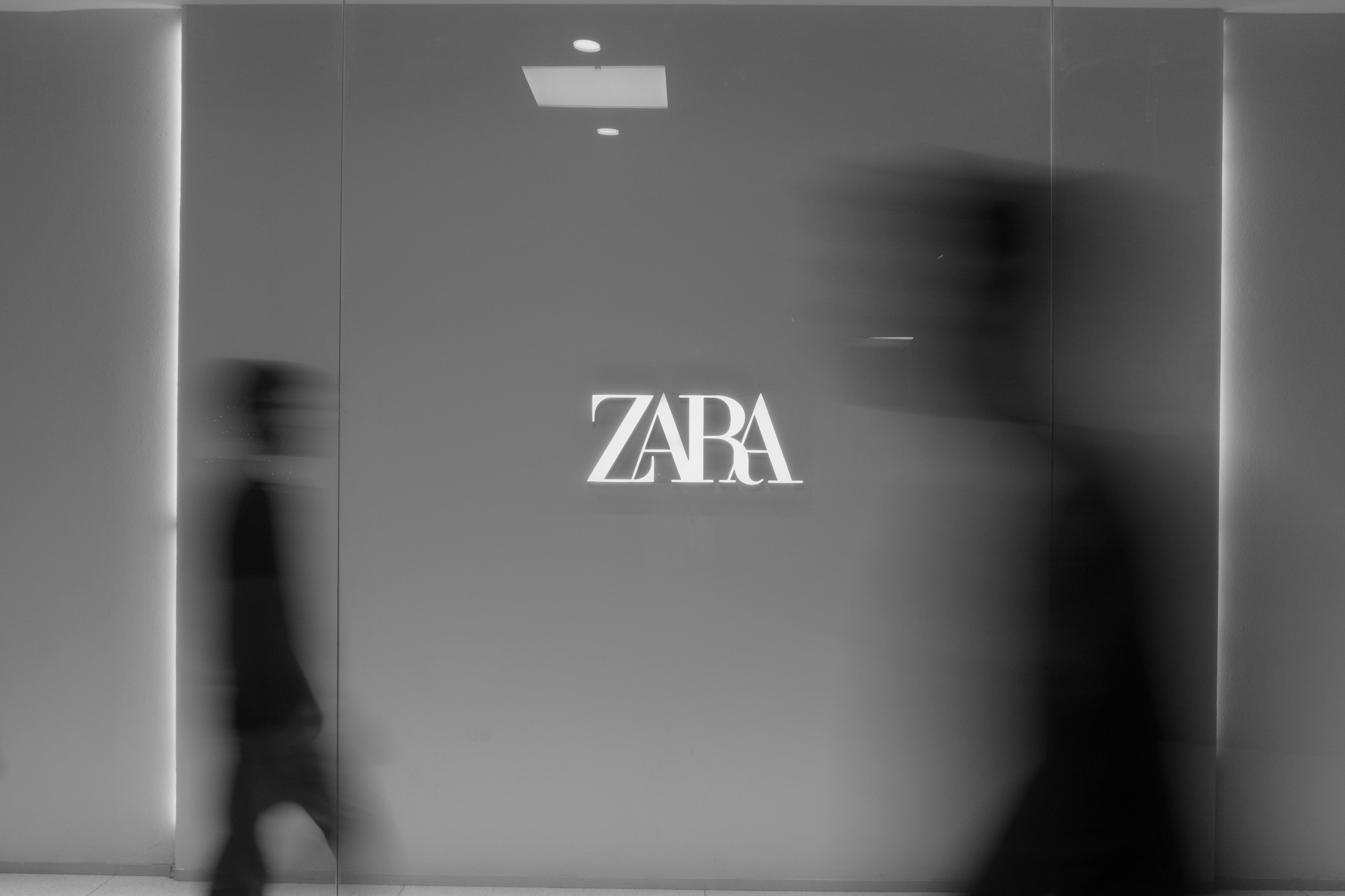 Blurred figures walk past a zaba logo on a wall.