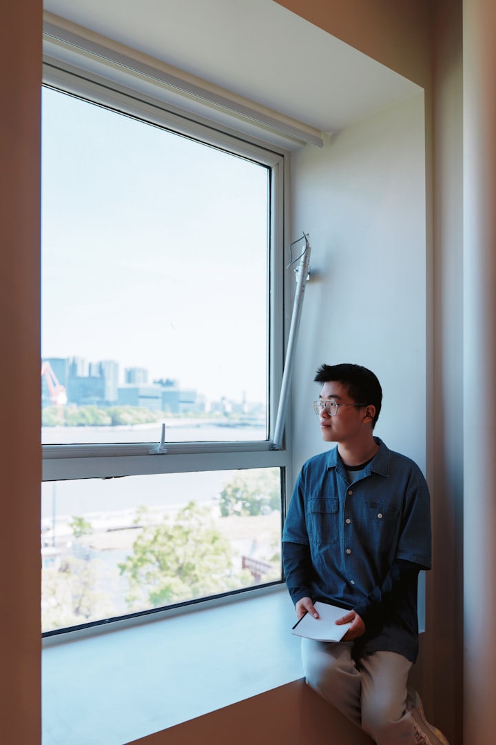 A young person sits by a window, looking out at a city view, anonymized profile framing