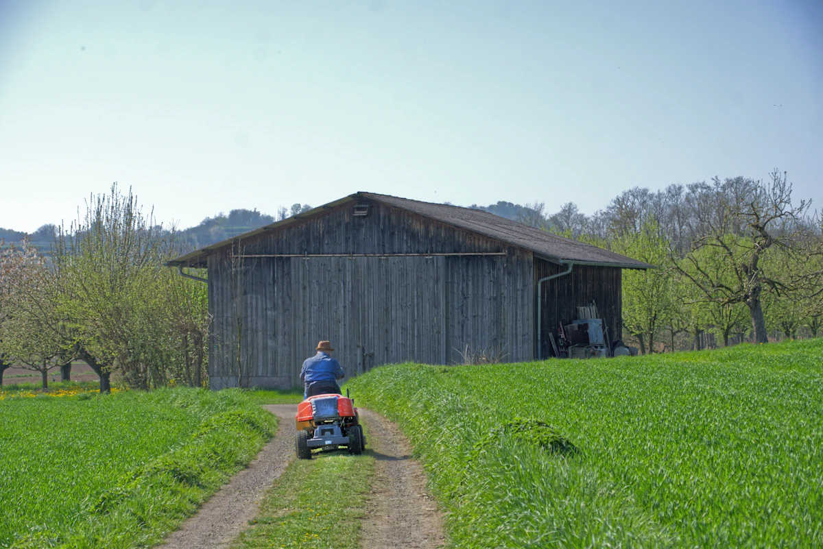 Man riding a mower on a dirt path near a barn