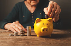 Elderly hands depositing coins into a yellow piggy bank.