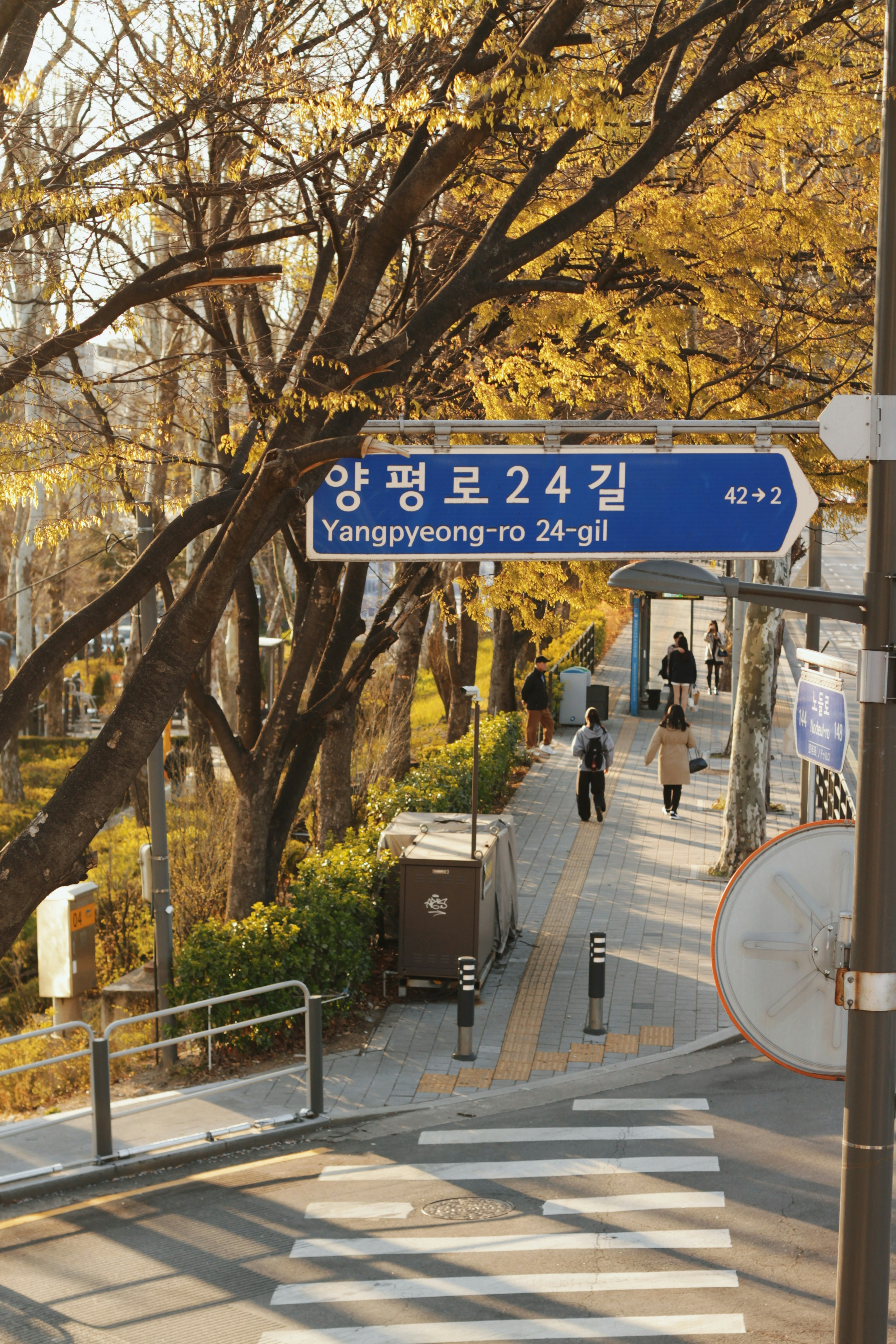 Placa de rua com texto coreano e pessoas andando na calçada.