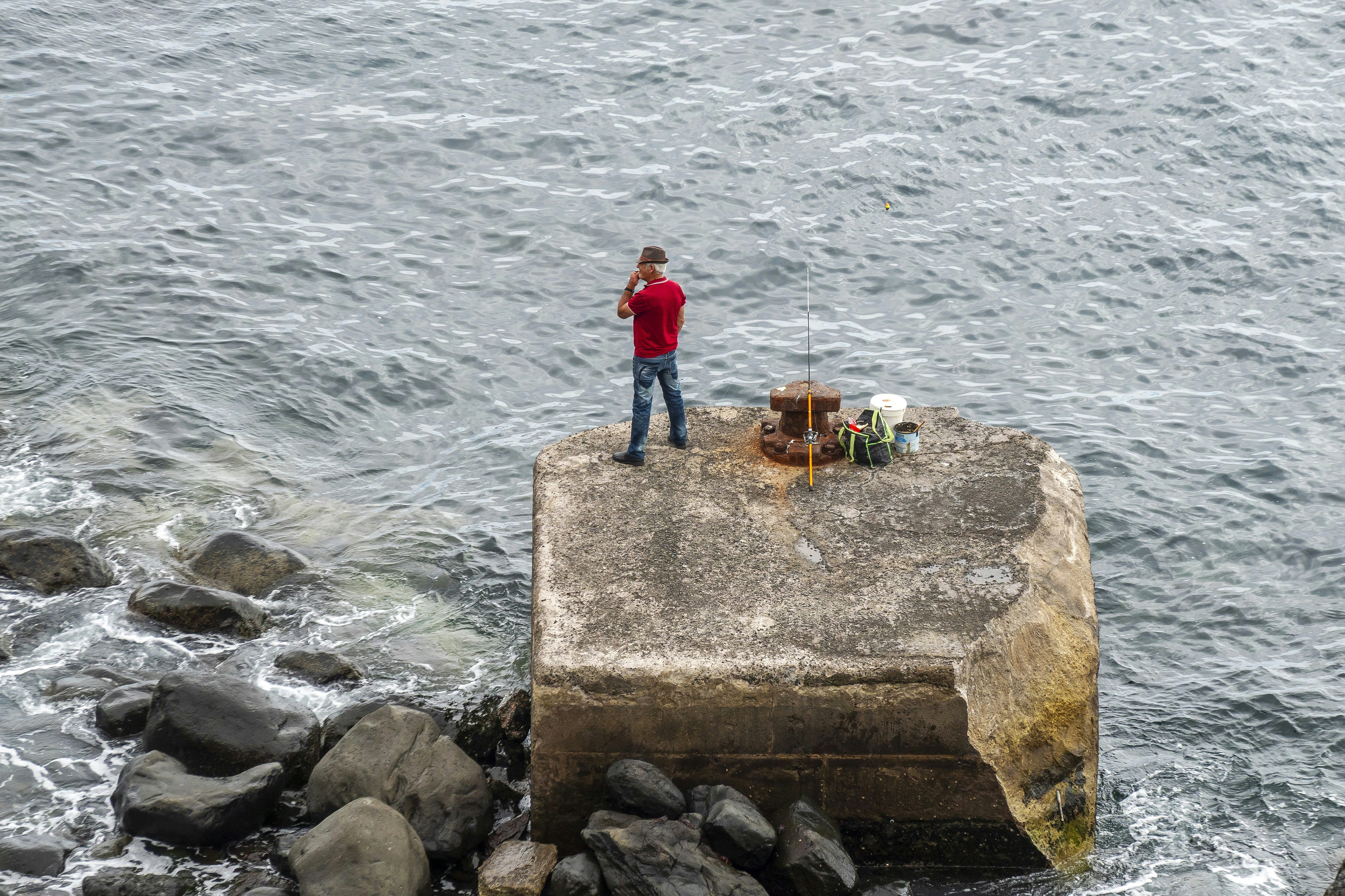 Man in red shirt stands on concrete block by ocean.