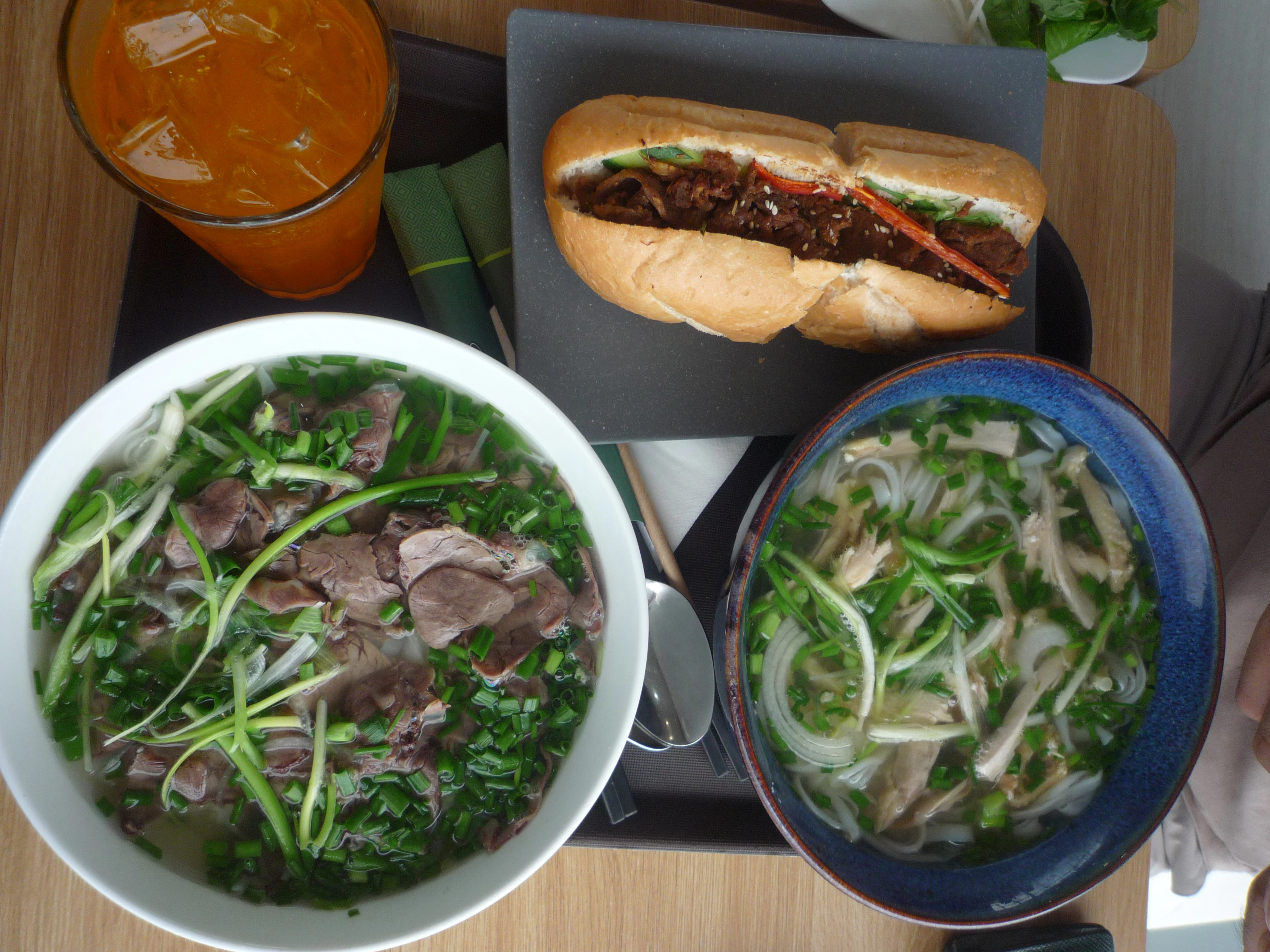 A Vietnamese meal seen from above — two bowls of phở with sliced beef, spring onion and coriander, a filled bánh mì baguette and a glass of iced orange juice