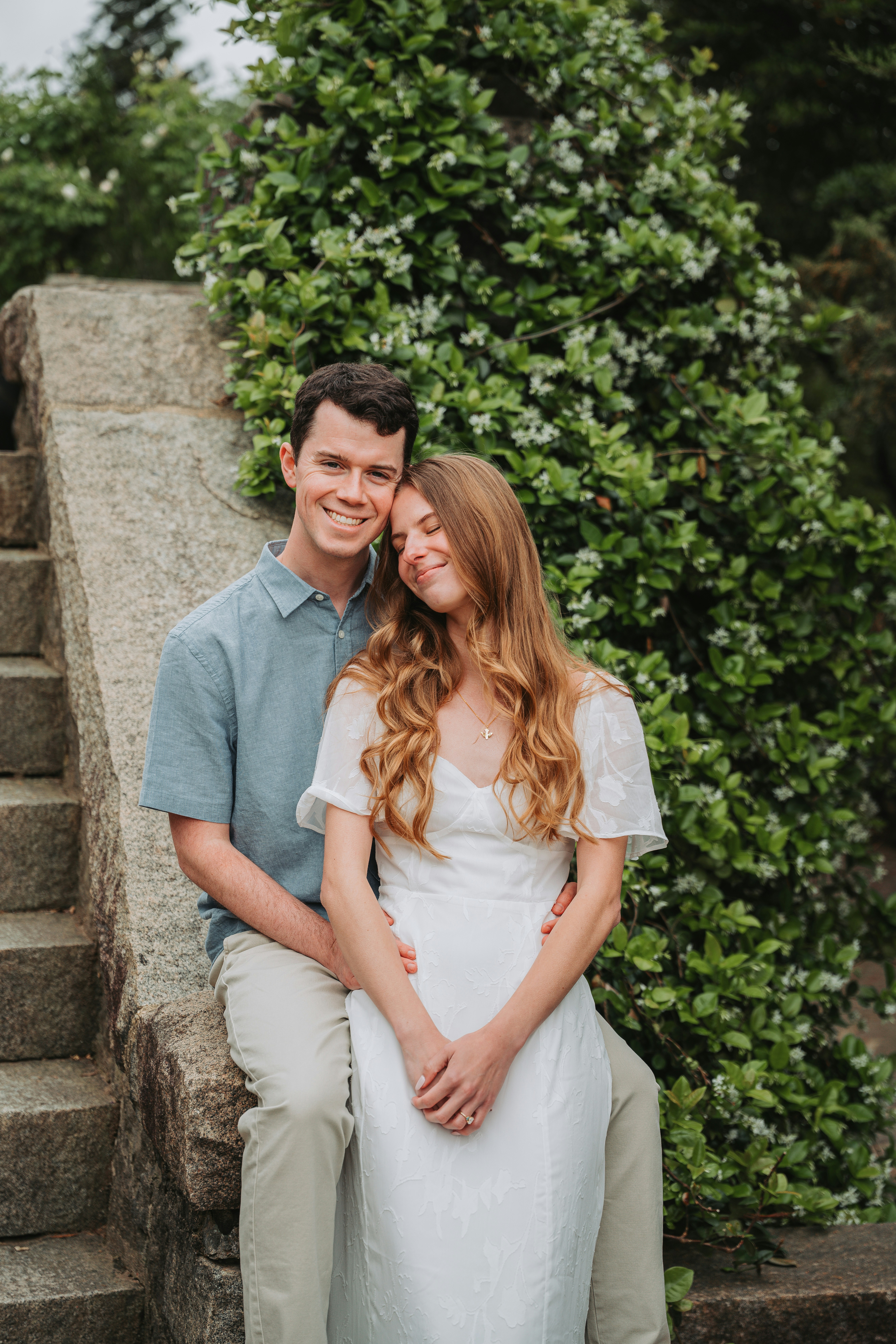 Um casal sorridente está sentado em degraus de pedra, cercado por vegetação.