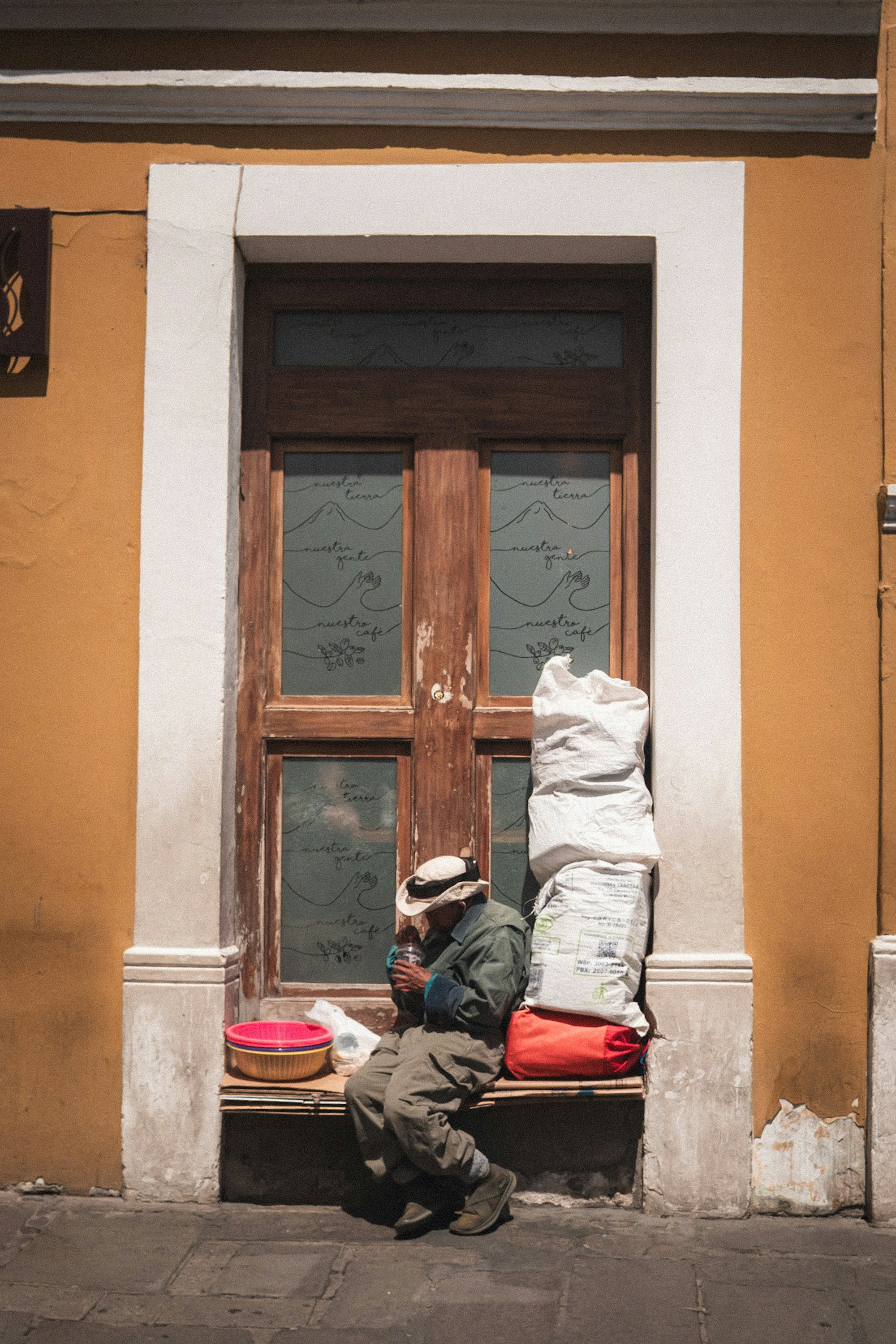 Un vieil homme assis près d’une porte en bois avec des sacs empilés.