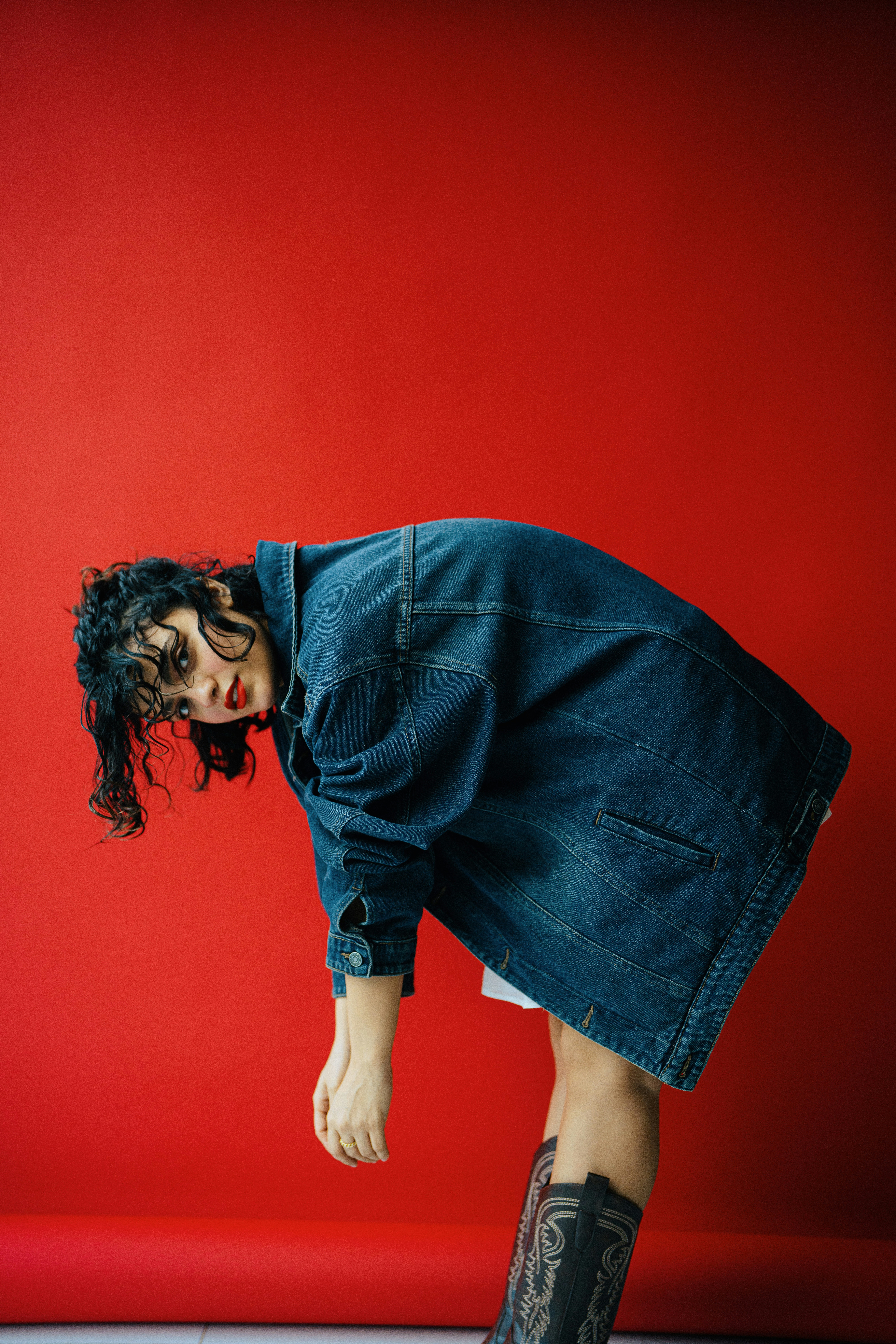 Woman in denim jacket bends over red backdrop