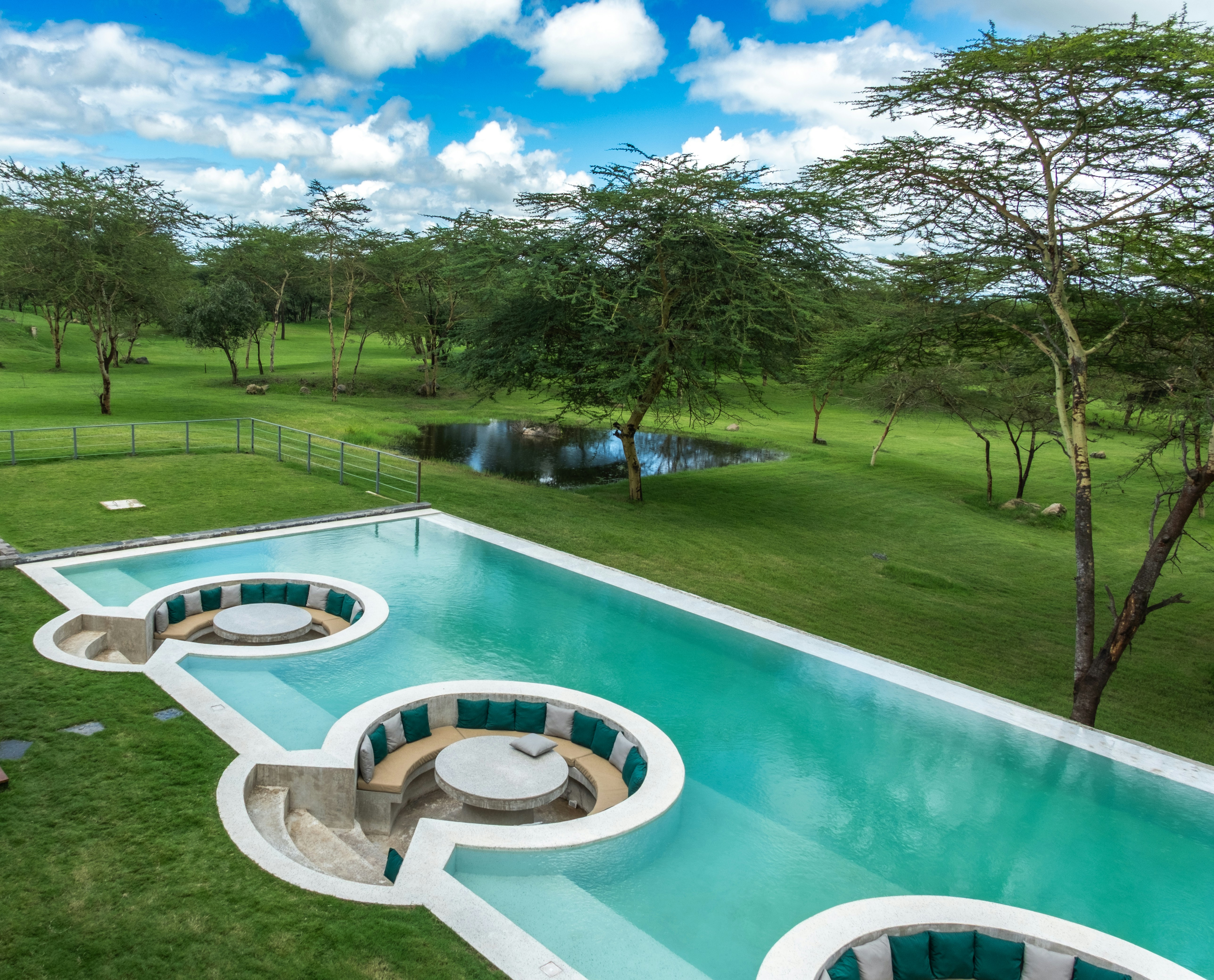 High-angle shot of a modern turquoise swimming pool featuring circular sunken lounge areas and stone tables, surrounded by green lawns and acacia trees.