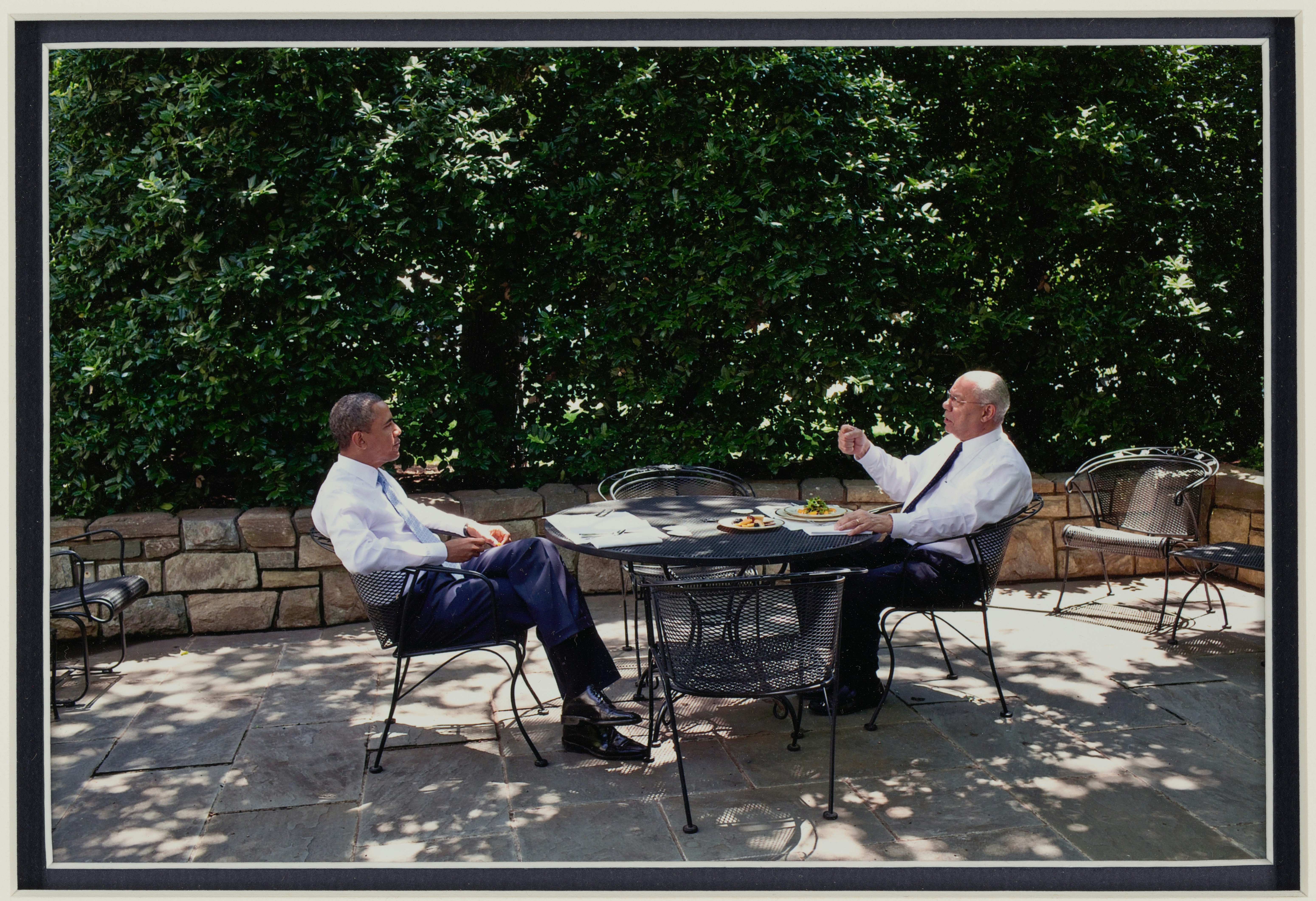 Deux hommes en costume discutant dehors à une table.