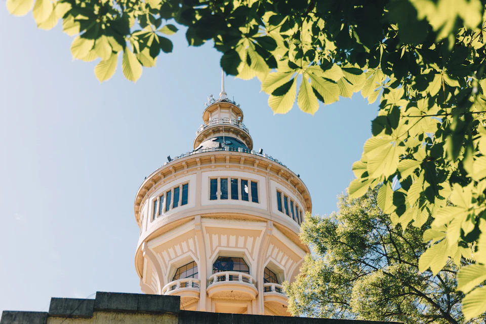 The image depicts a large, white building with a round tower and multiple levels, situated on a street corner. The building is surrounded by lush green trees, creating a serene atmosphere. The perspective of the photo is from below, looking up at the towering structure against a clear blue sky.