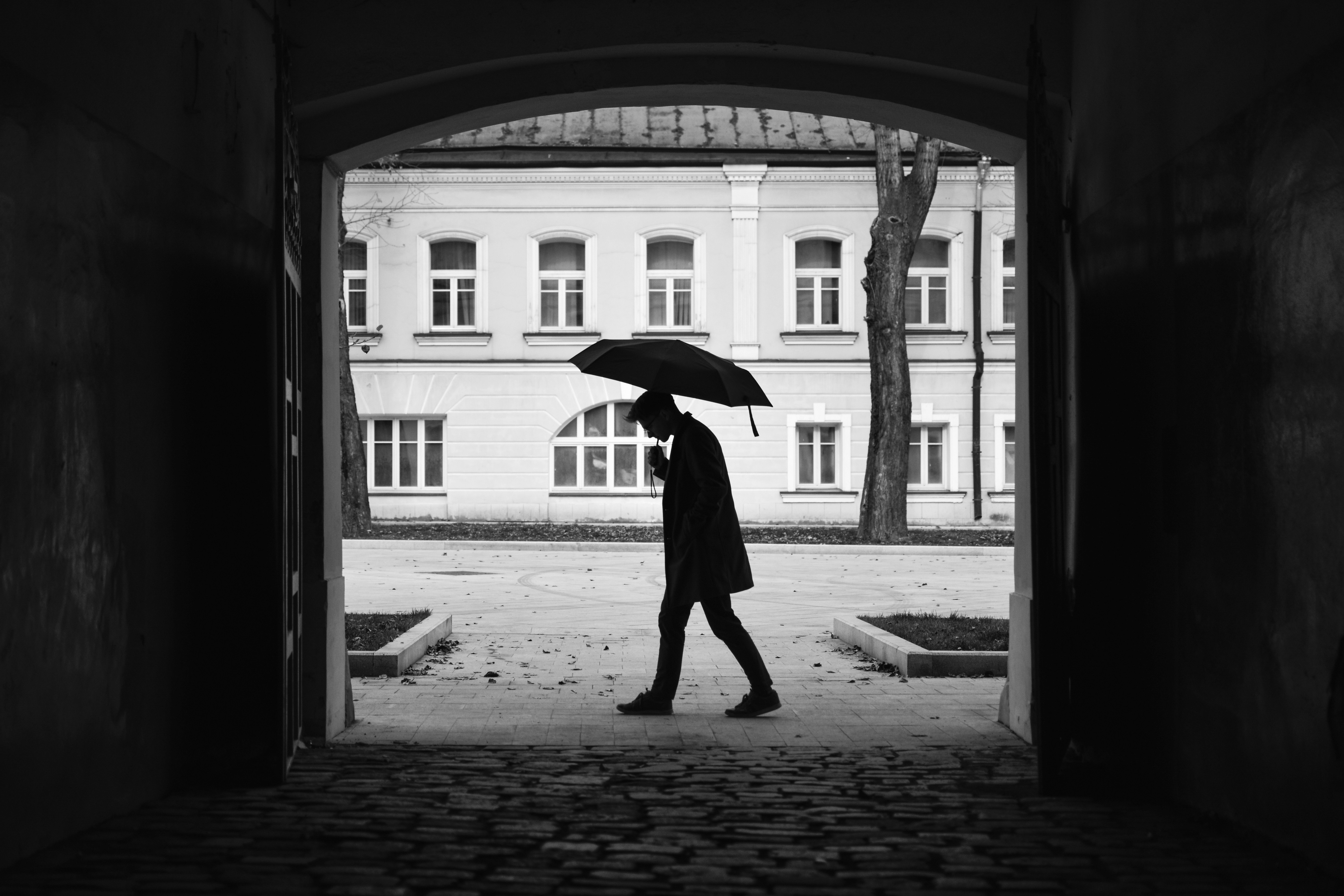 A cinematic black and white shot of a lone man walking with an umbrella through a stone archway. The composition uses natural framing to highlight the silhouette against a classic architectural backdrop, evoking a sense of urban solitude and moody atmosphere.