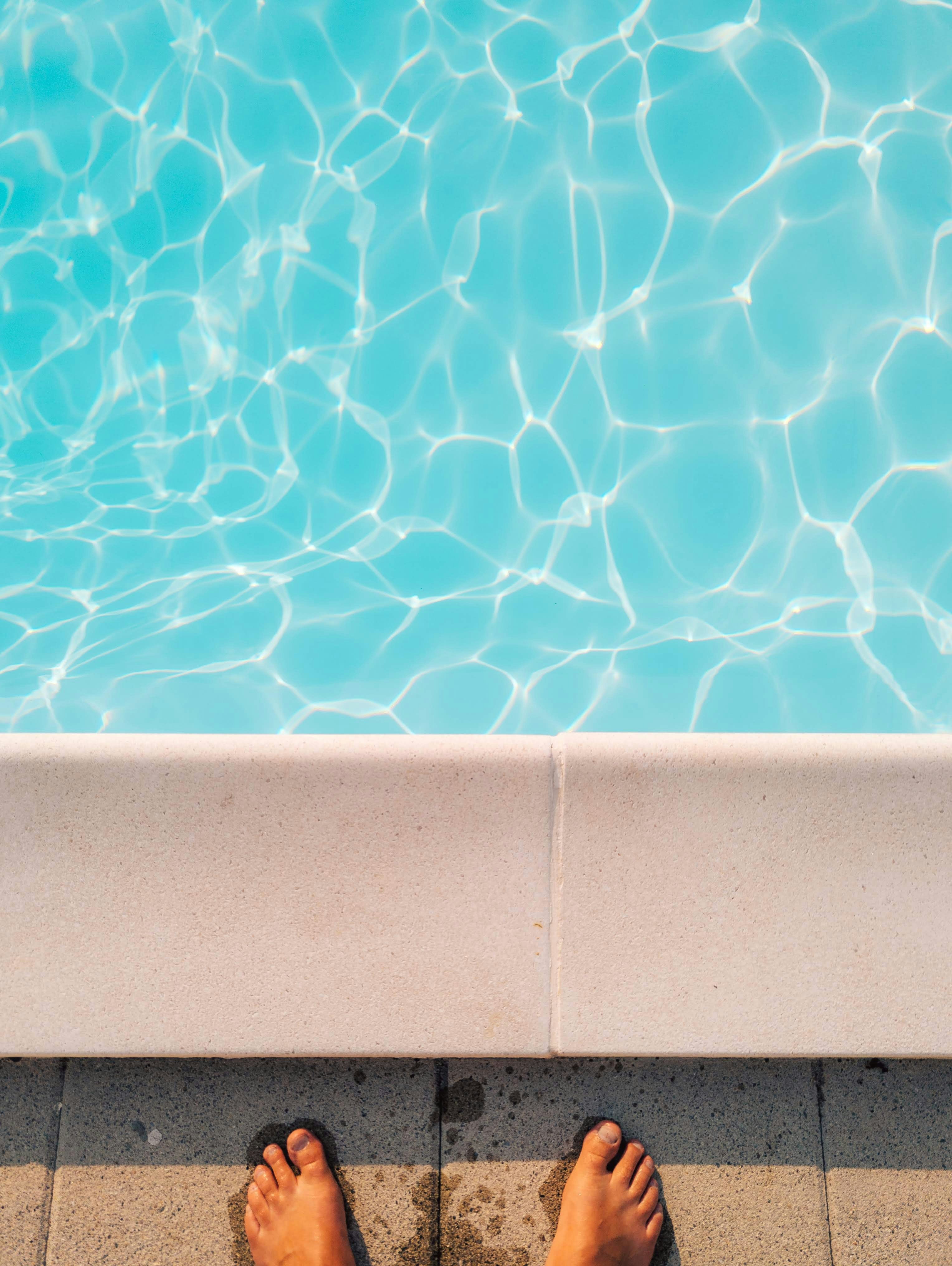 Bare feet on pavement by a bright blue swimming pool.