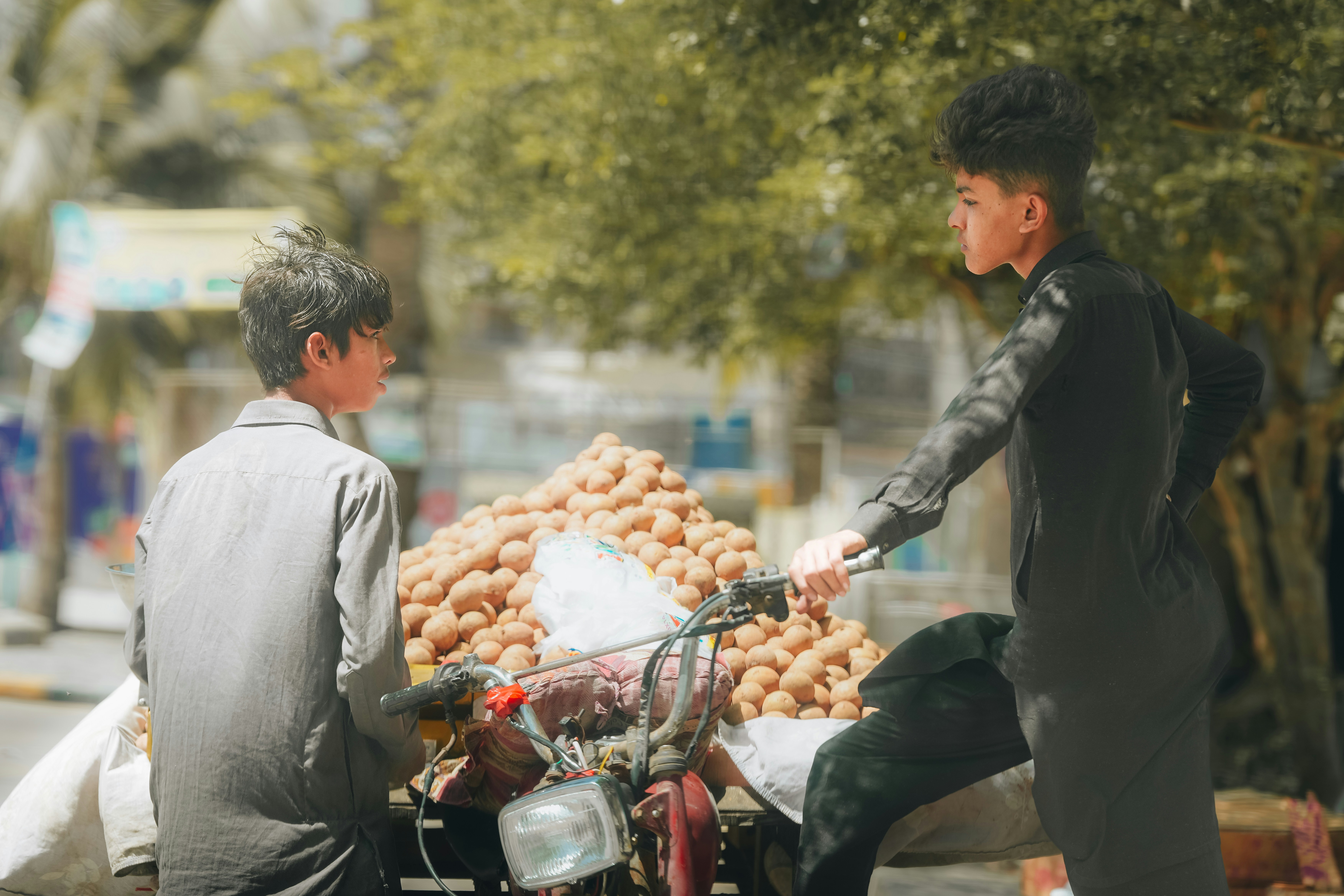 Two boys interact at a fruit stand with a motorcycle.