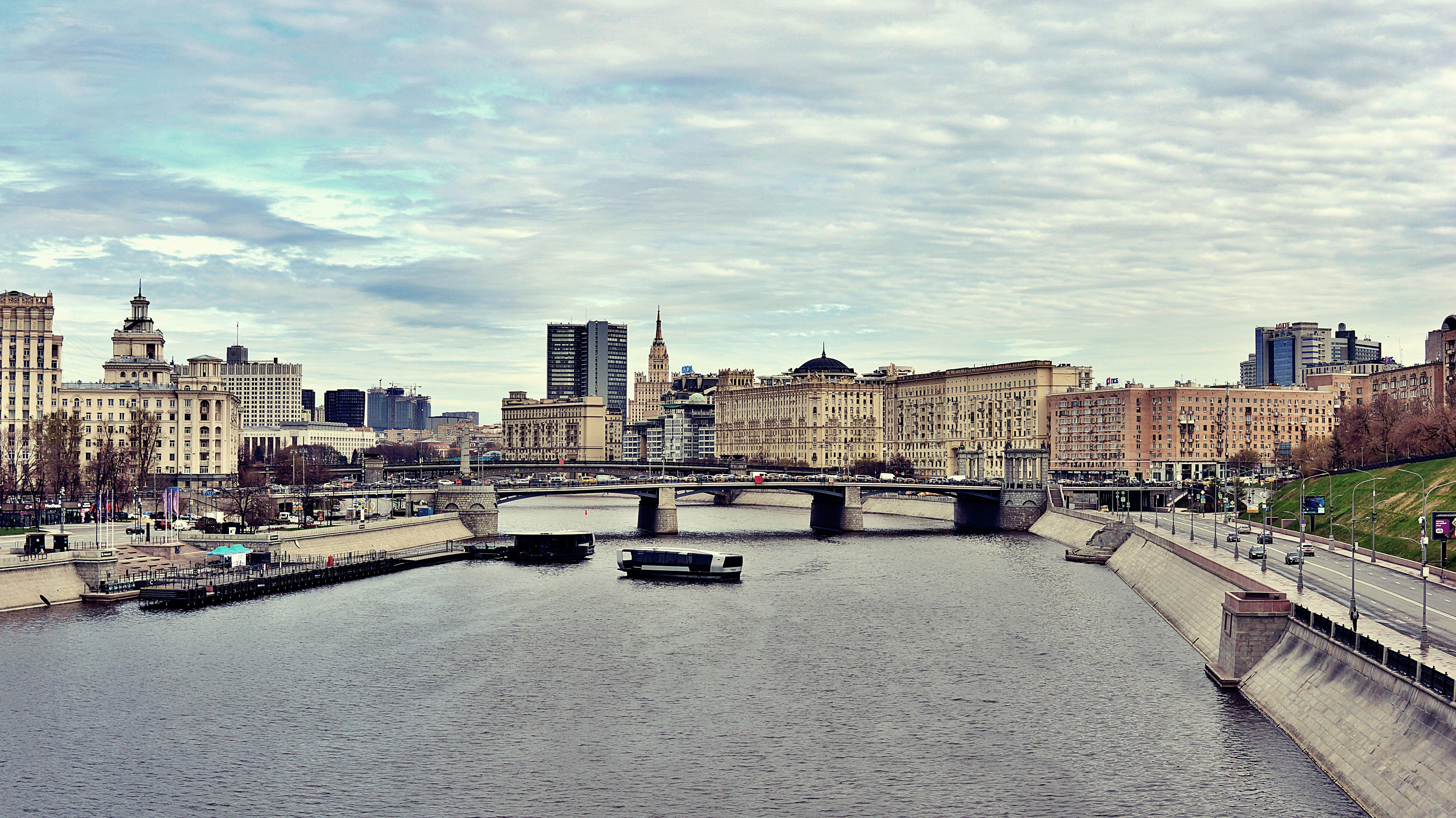 Cityscape with river, bridge, and buildings under cloudy sky.