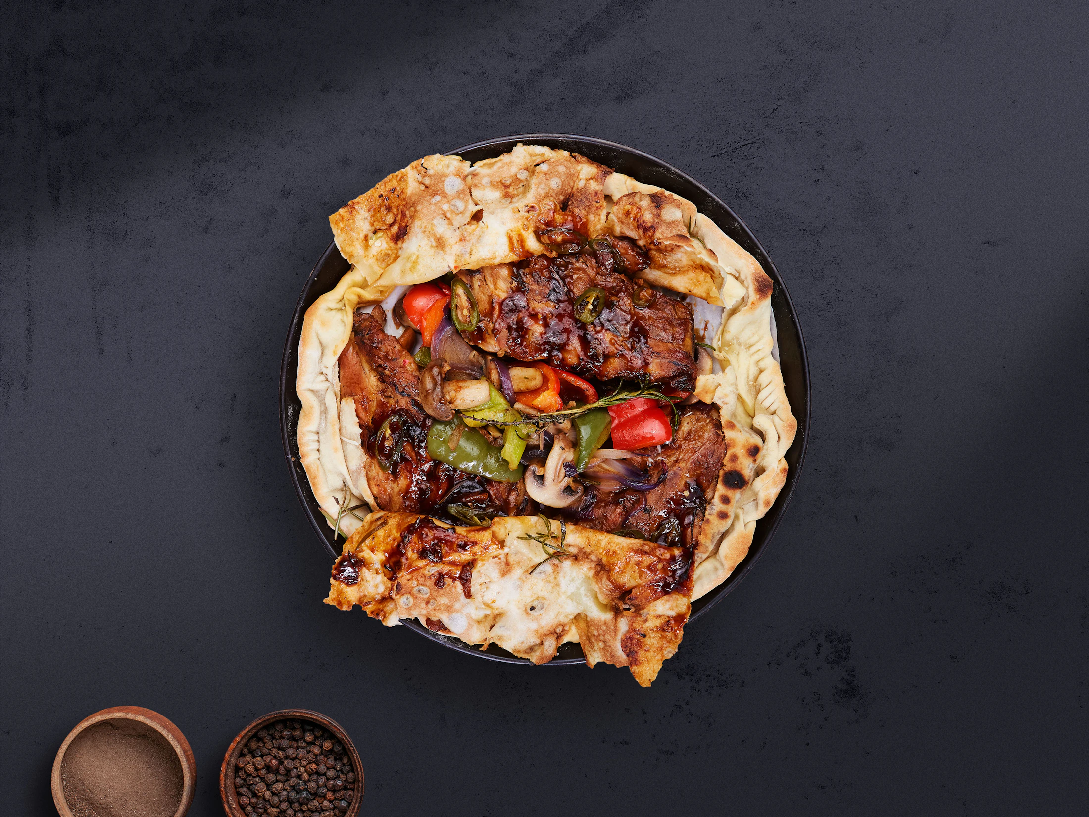 Flatbread topped with grilled meat and roasted vegetables on dark slate