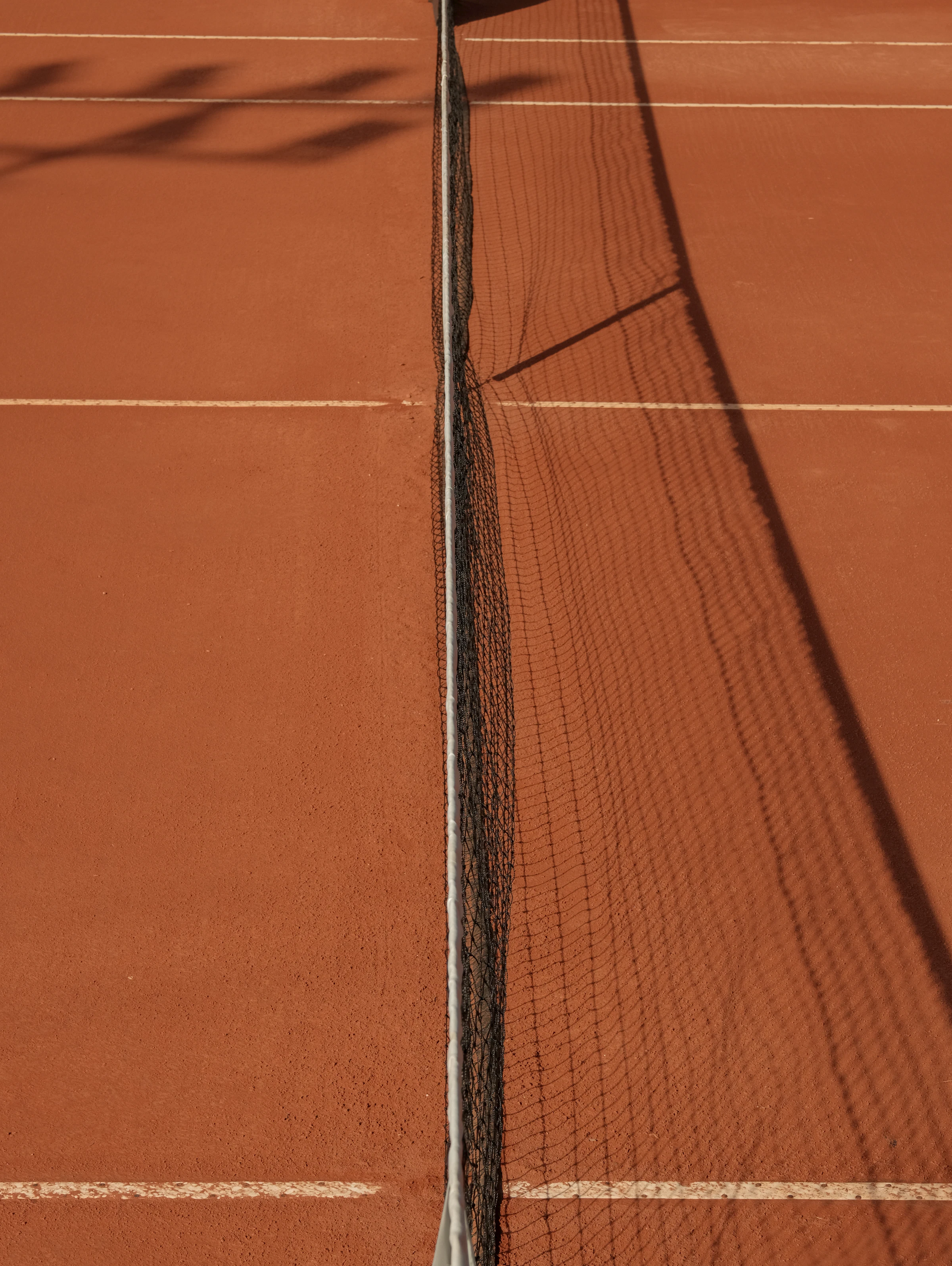 A tennis net casting a long shadow across a red clay court at golden hour.