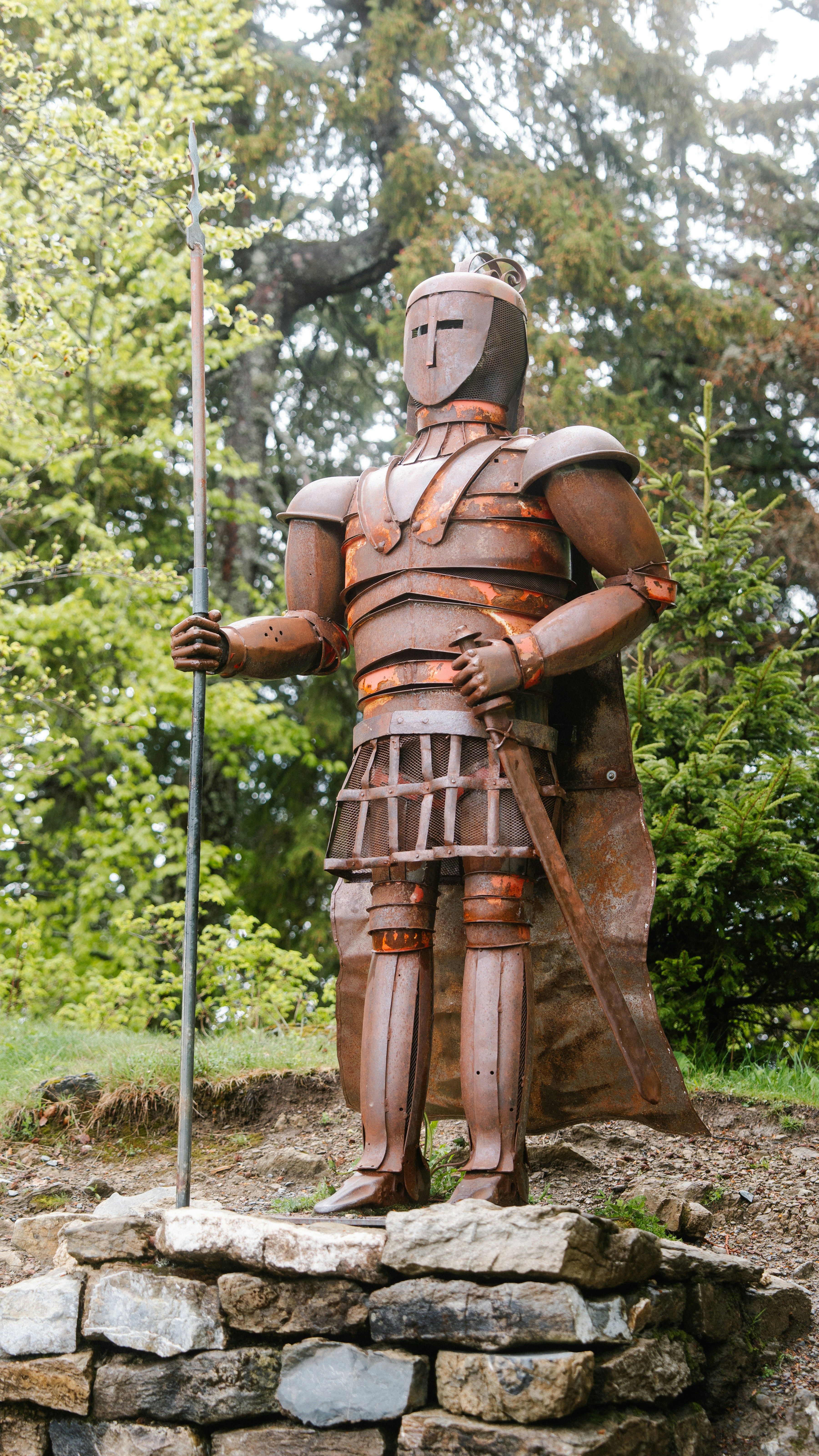 Rusty knight statue holding spear and sword outdoors.