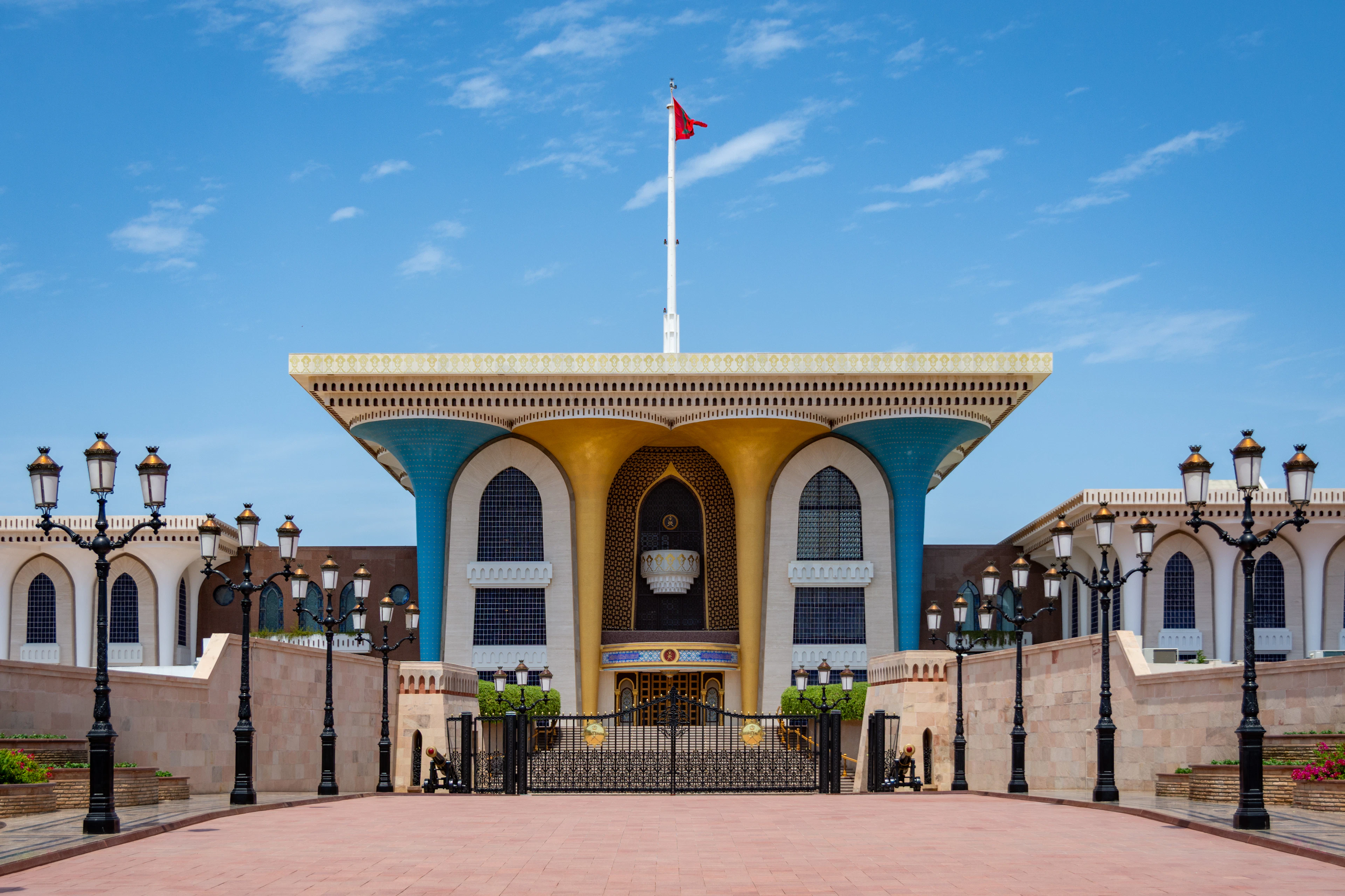The blue and gold flared columns of Al Alam Palace in Muscat under a clear sky, its ceremonial forecourt empty between two rows of black lamp posts.