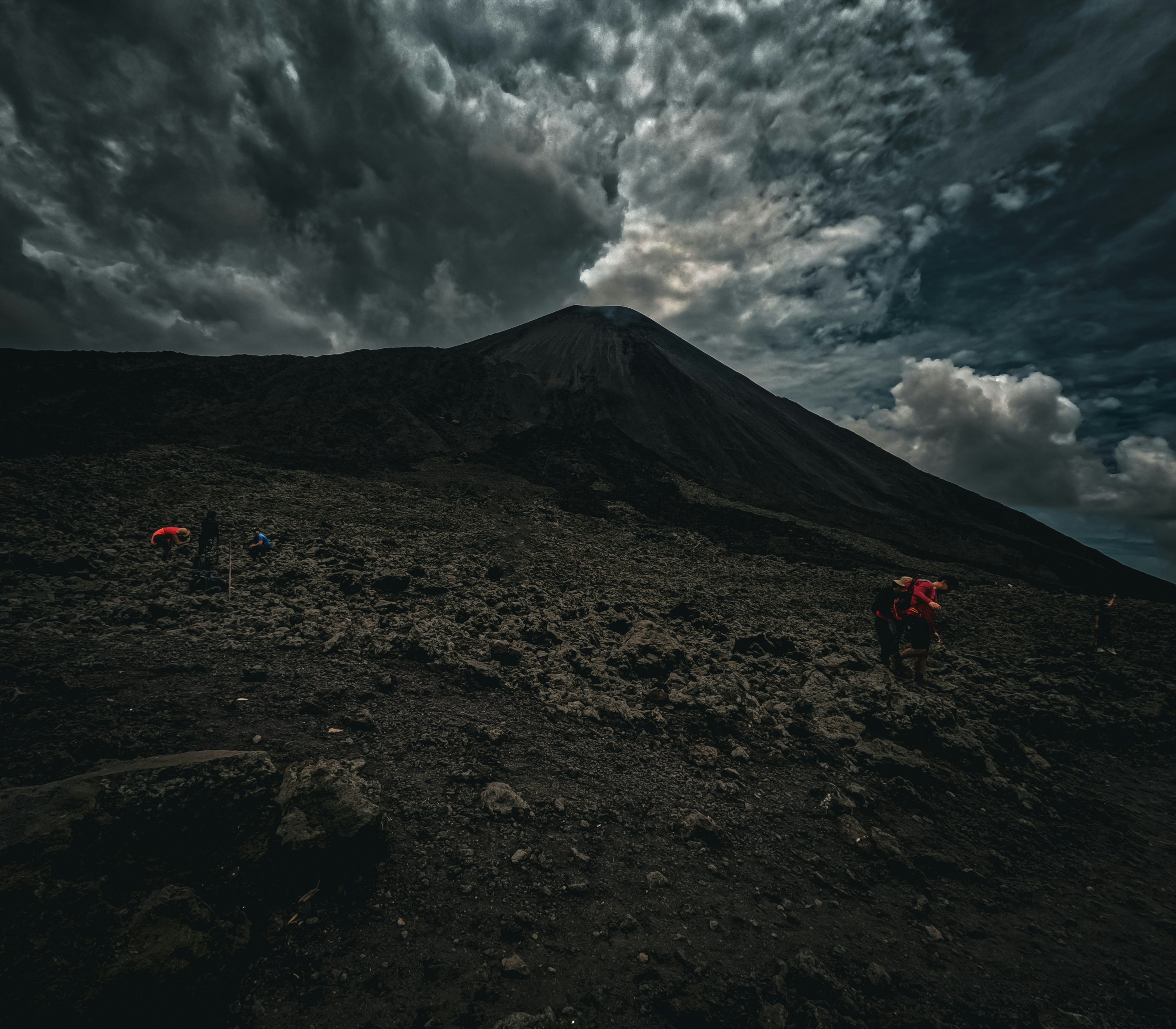 Volcanic slope at dusk with climbers scattering across rugged terrain; dramatic clouds loom above a dark peak.