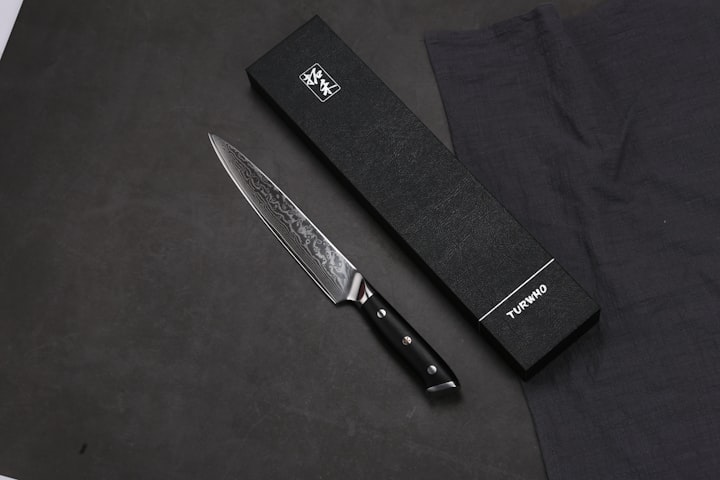 A Damascus chef's knife on a dark surface, the laminated steel pattern echoing the heavy heel of a deba filleting blade