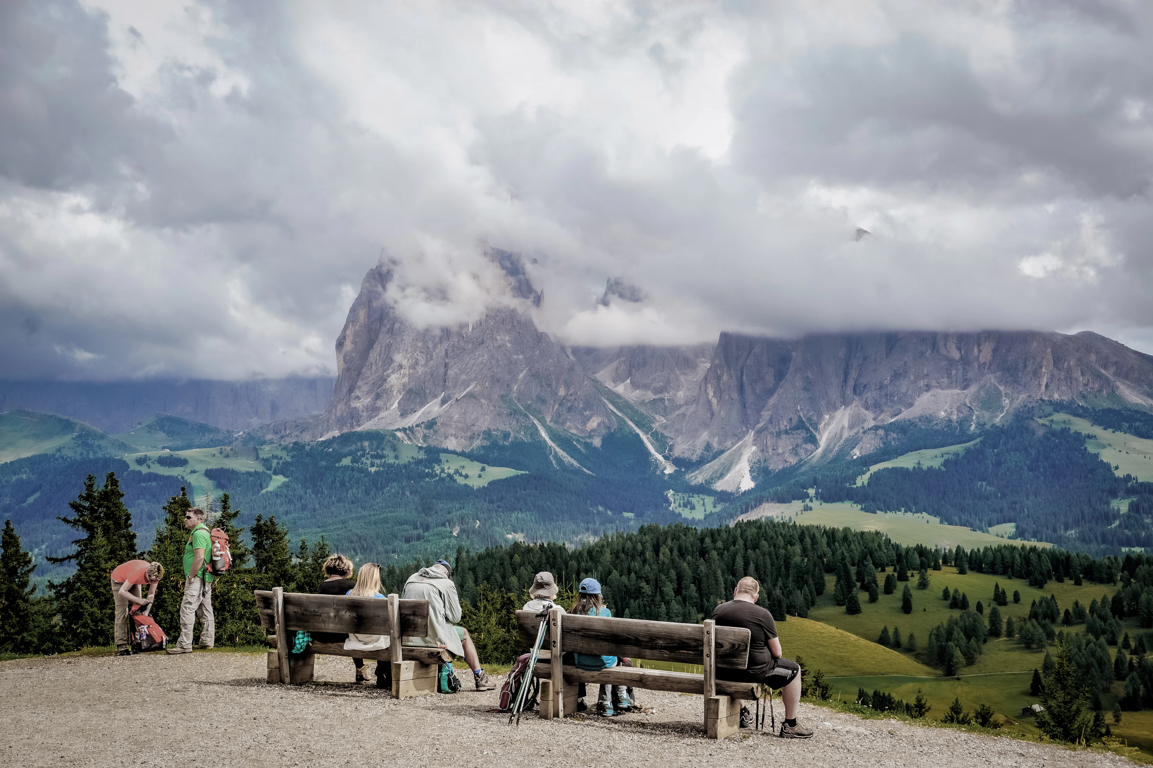 People resting on benches at a viewpoint facing the Dolomites, Alpe di Siusi