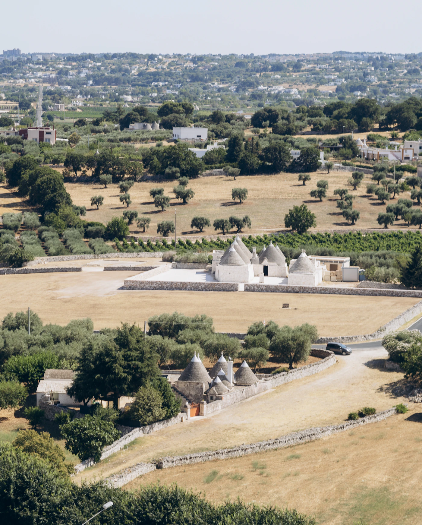 A traditional trulli house in the Itria Valley Puglia with a whitewashed conical roof rising above a rural stone wall with the green rolling countryside visible beyond in soft Italian light