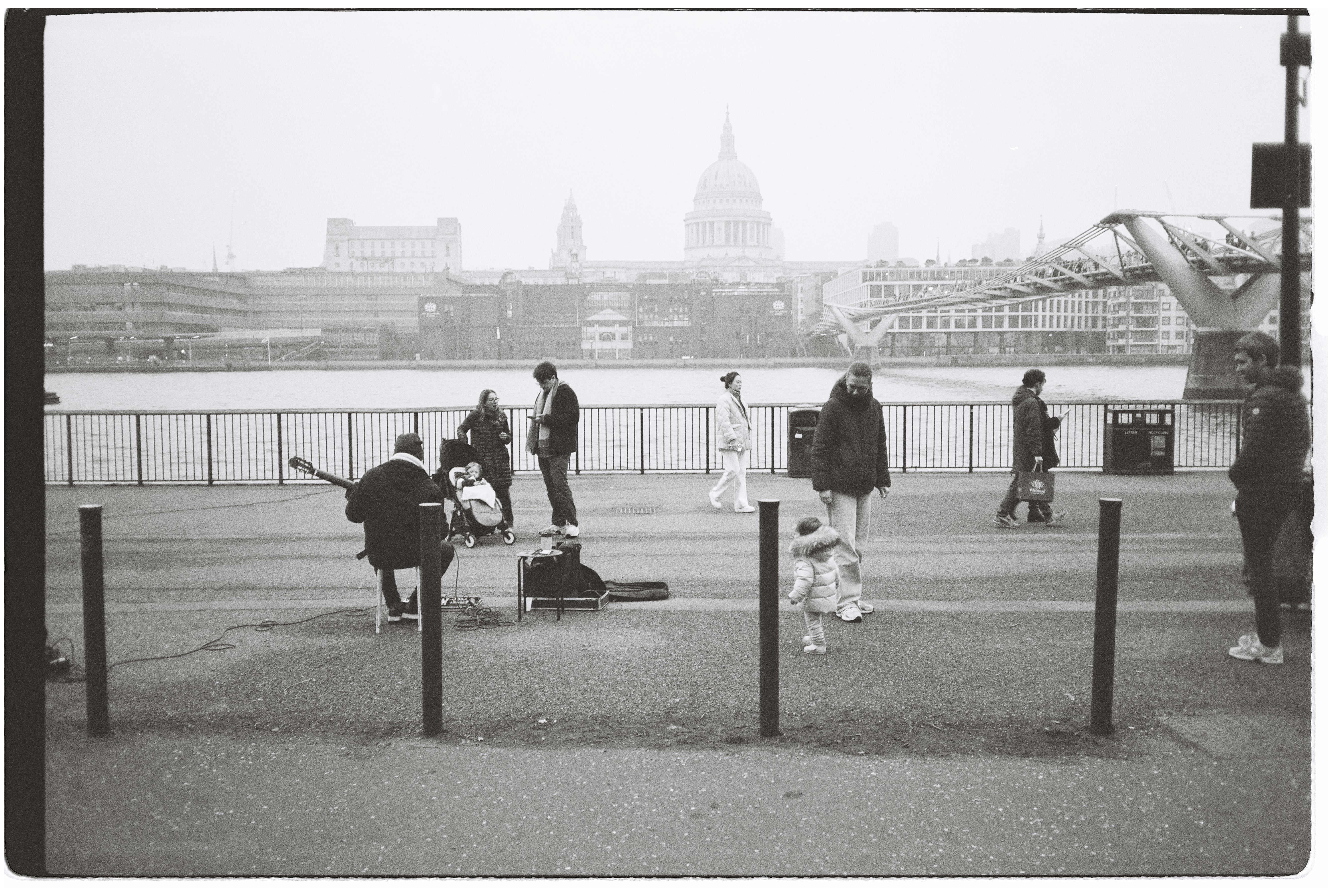 Musician plays guitar by river with city skyline background.
