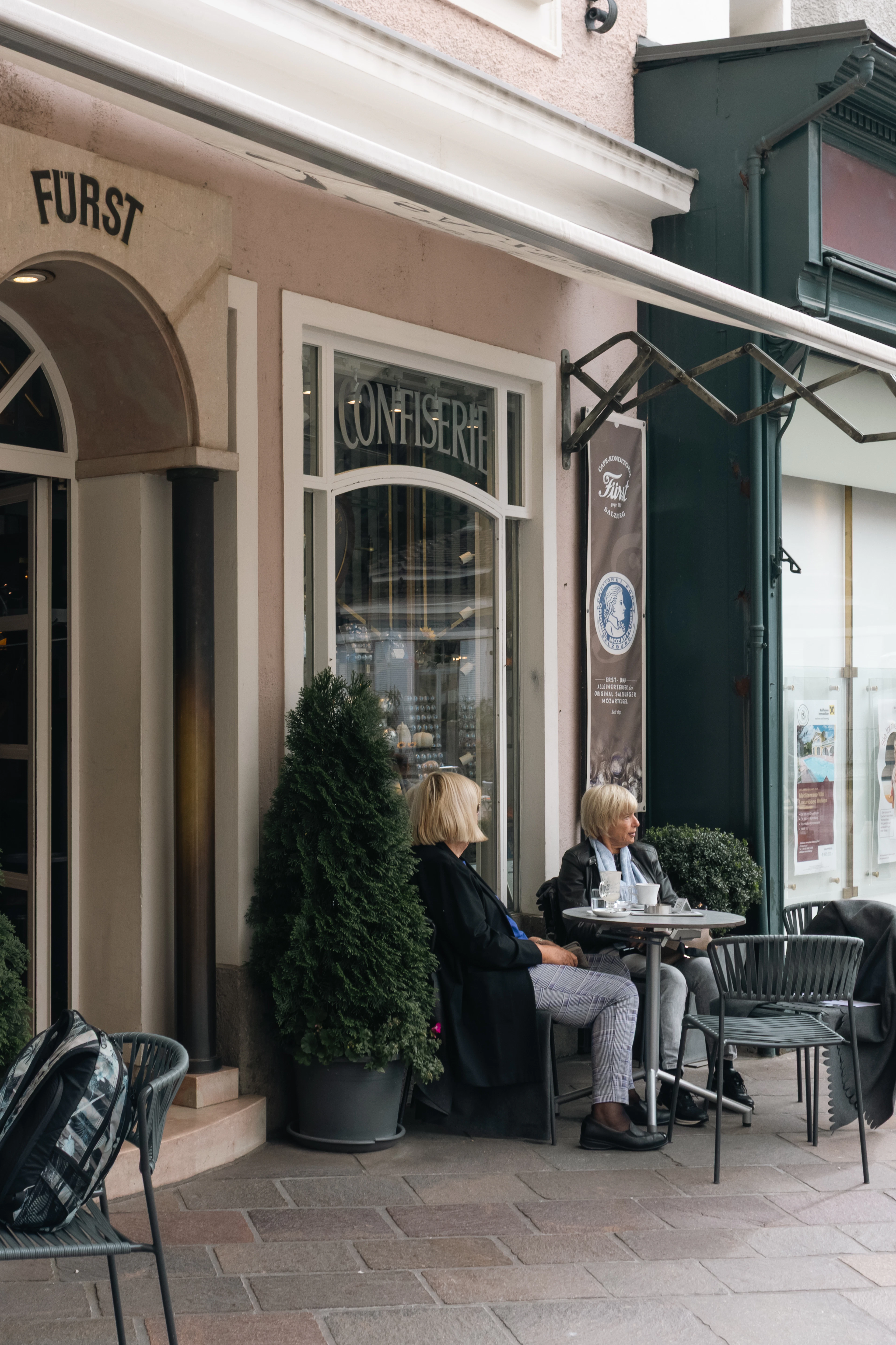 Two women share coffee at an outdoor table outside the Furst confectionery on a Salzburg street.
