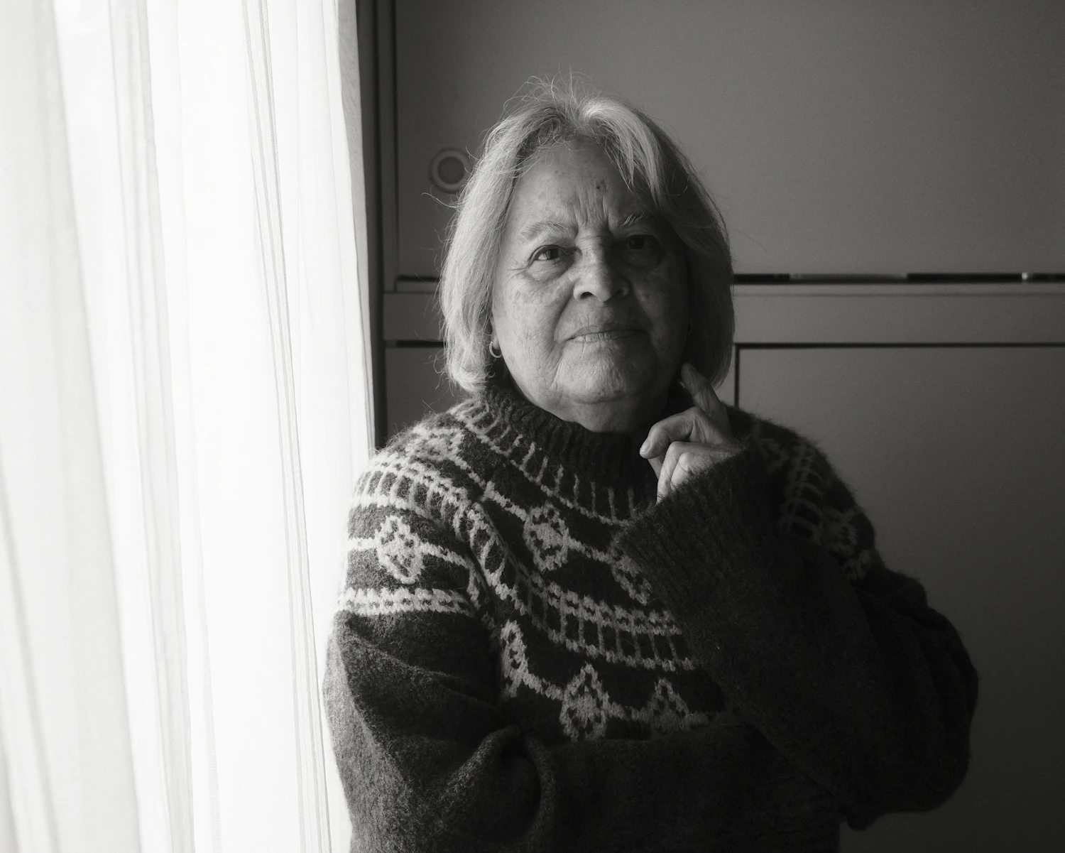 An older woman in a patterned knitted sweater standing by a window