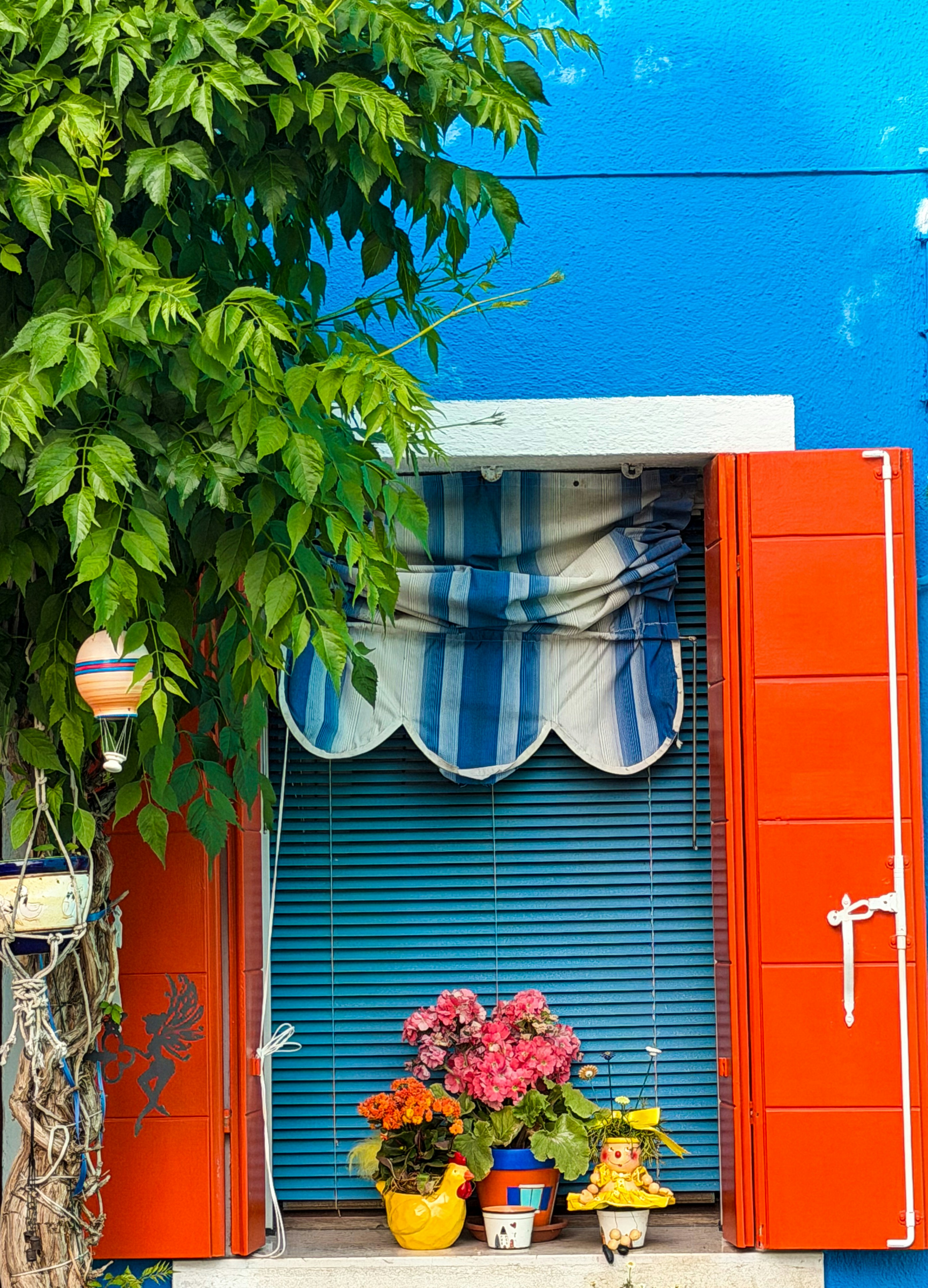 Des volets orange vif encadrent une fenêtre bleue ornée de fleurs en pot.