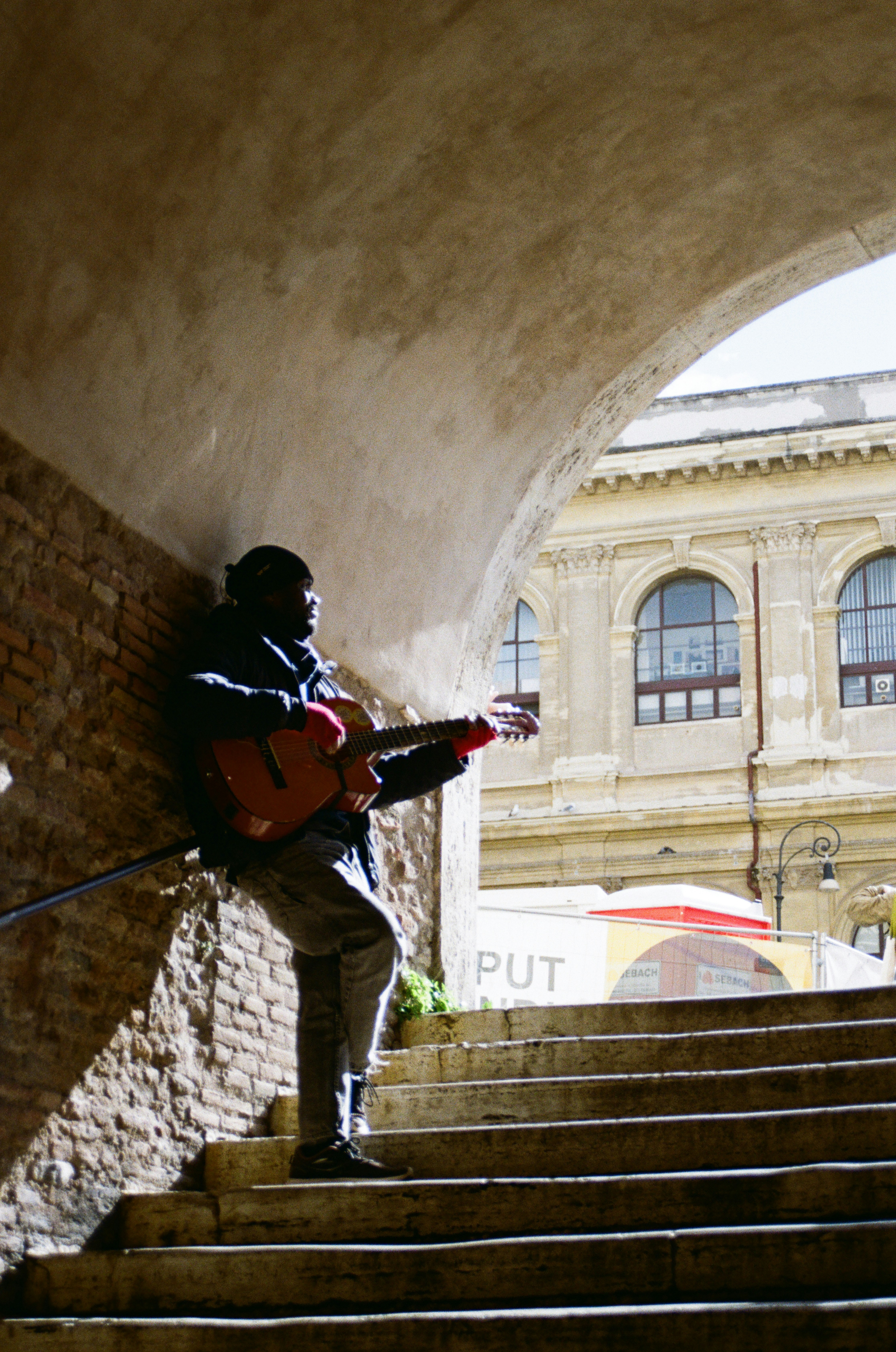 Man playing guitar on stone steps under archway.