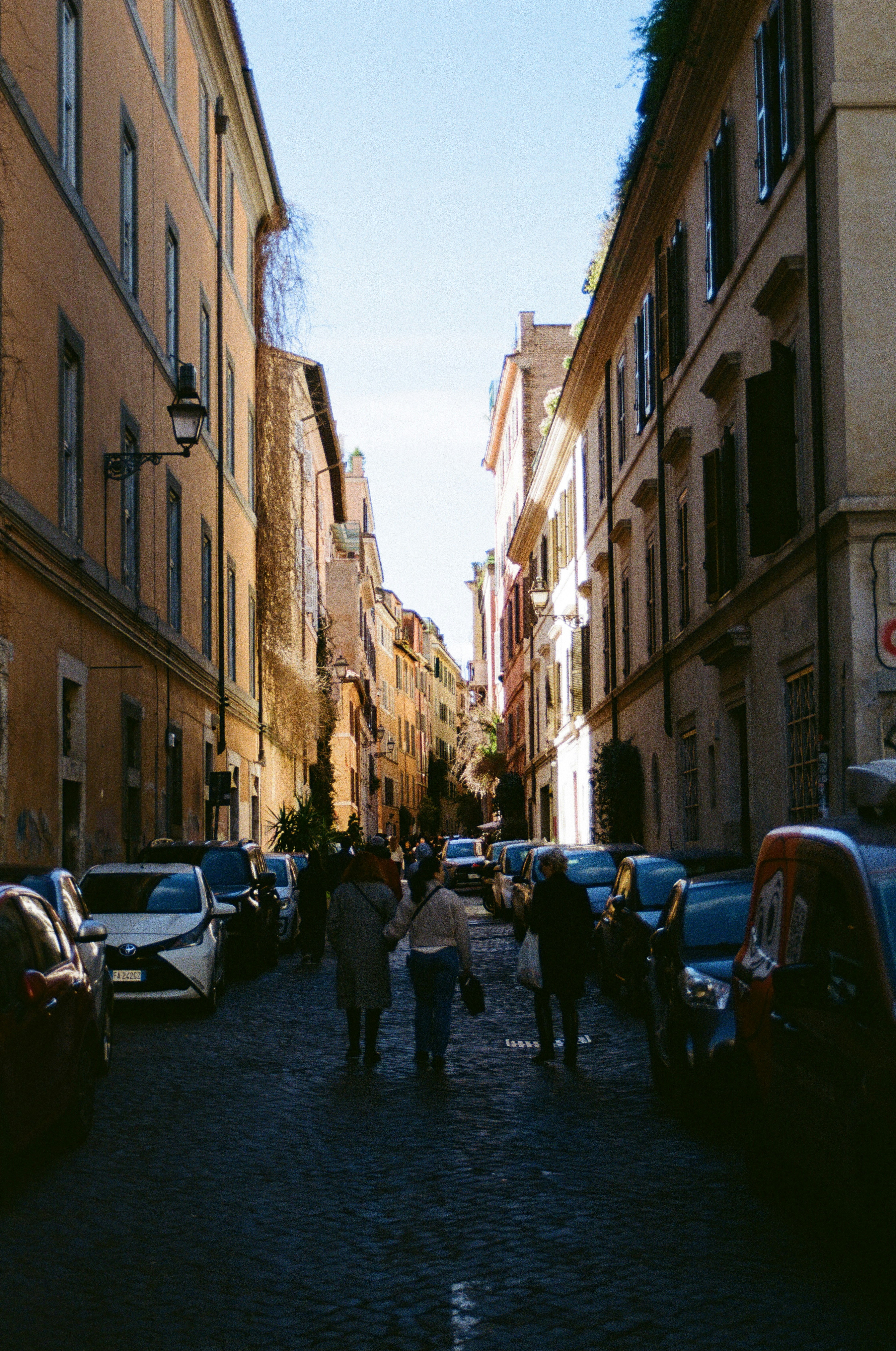 People walk down a narrow street lined with parked cars.