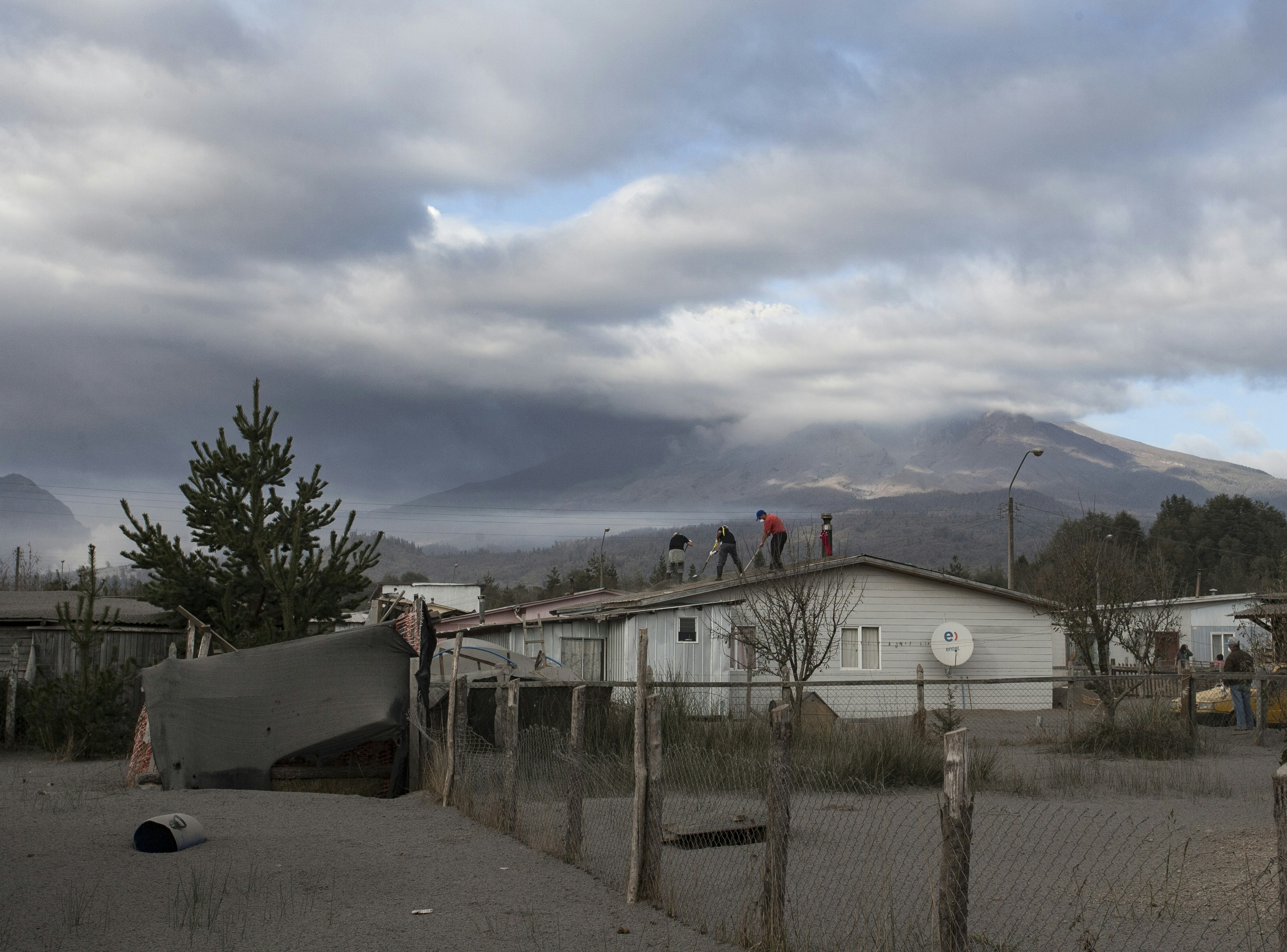 People on a house roof near a cloudy, ash-covered volcano