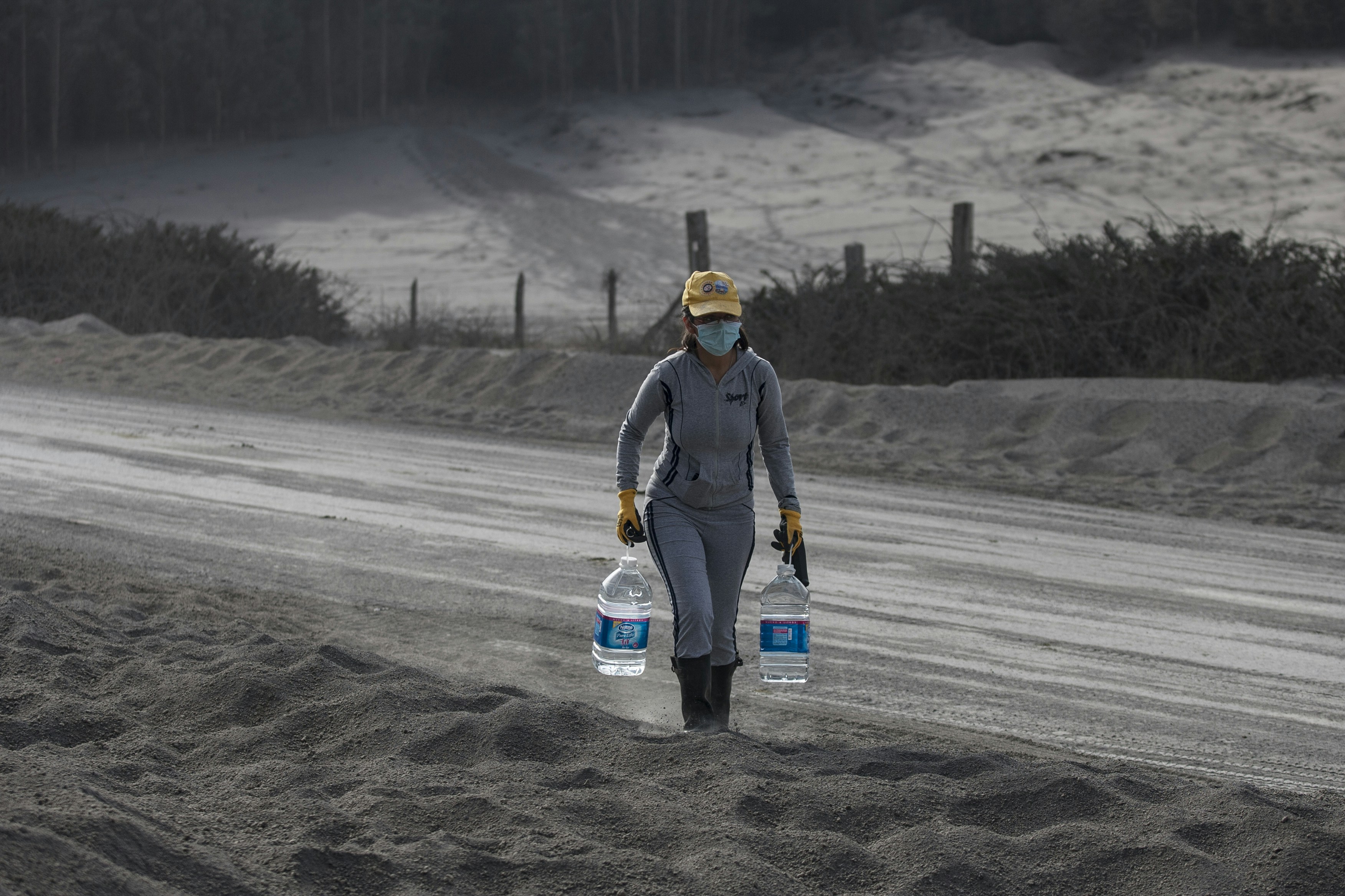 Person carries two large water bottles on ash-covered road