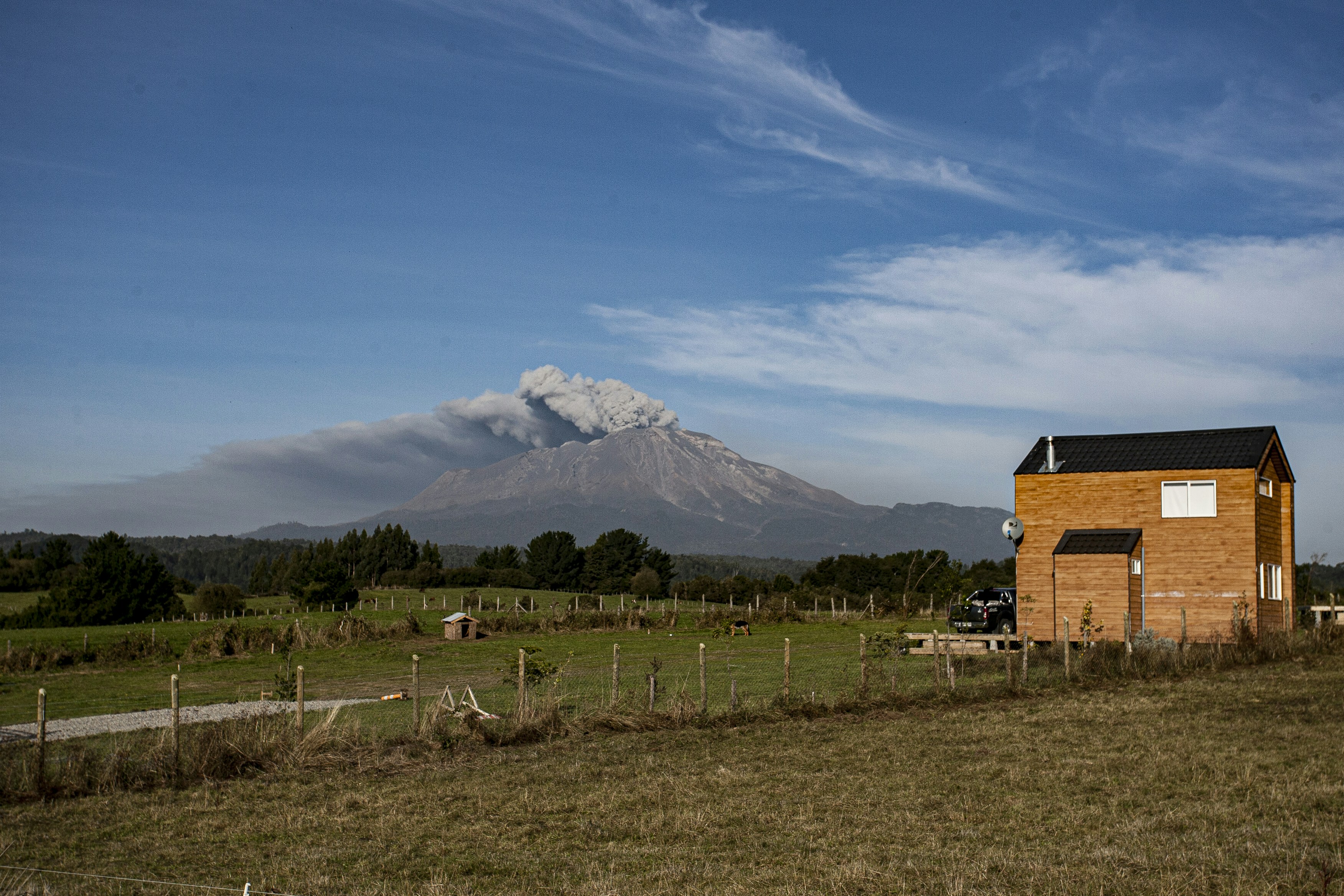 Volcano erupting with smoke over a rural landscape and house.