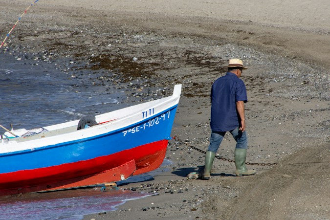 Man in a straw hat, jeans and mid-calf green rubber boots walking up a pebble beach, pulling a blue and red wooden boat ashore on a chain