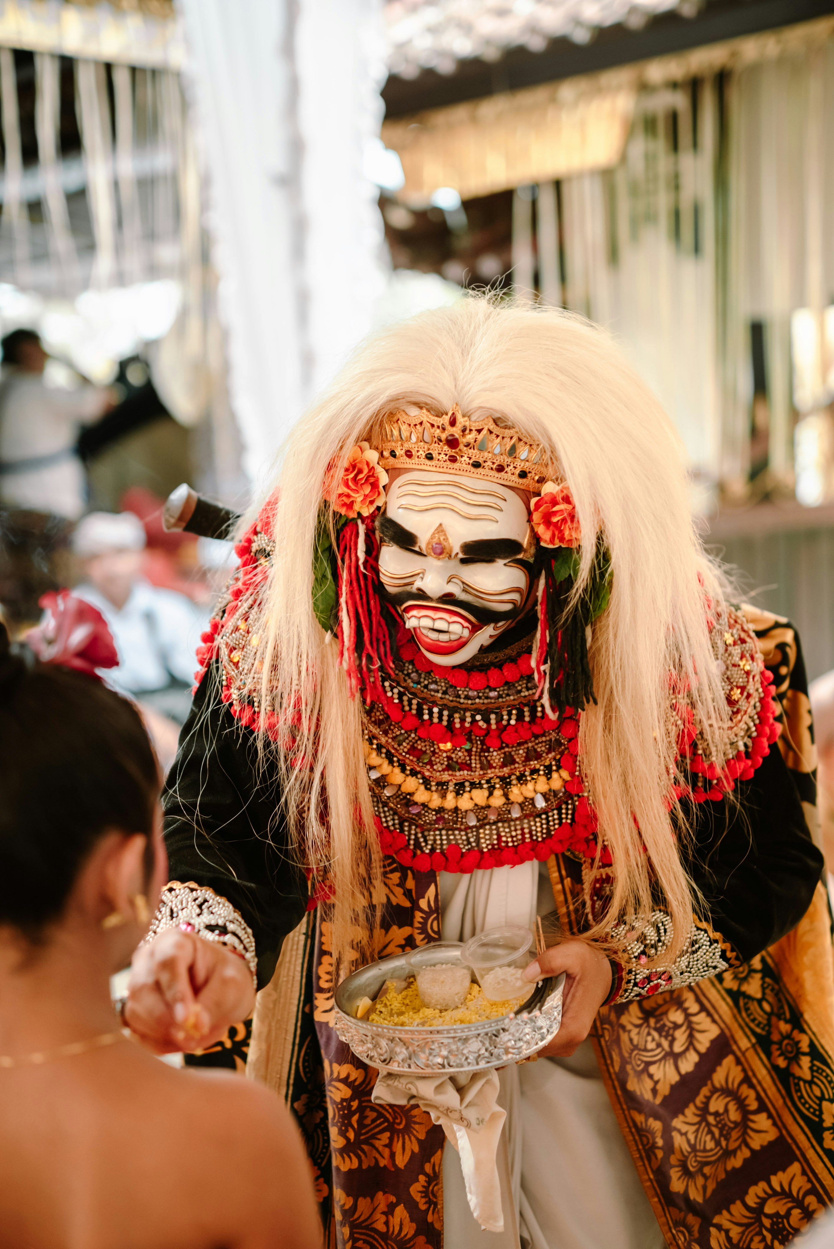 Man in elaborate mask offers food from a bowl.
