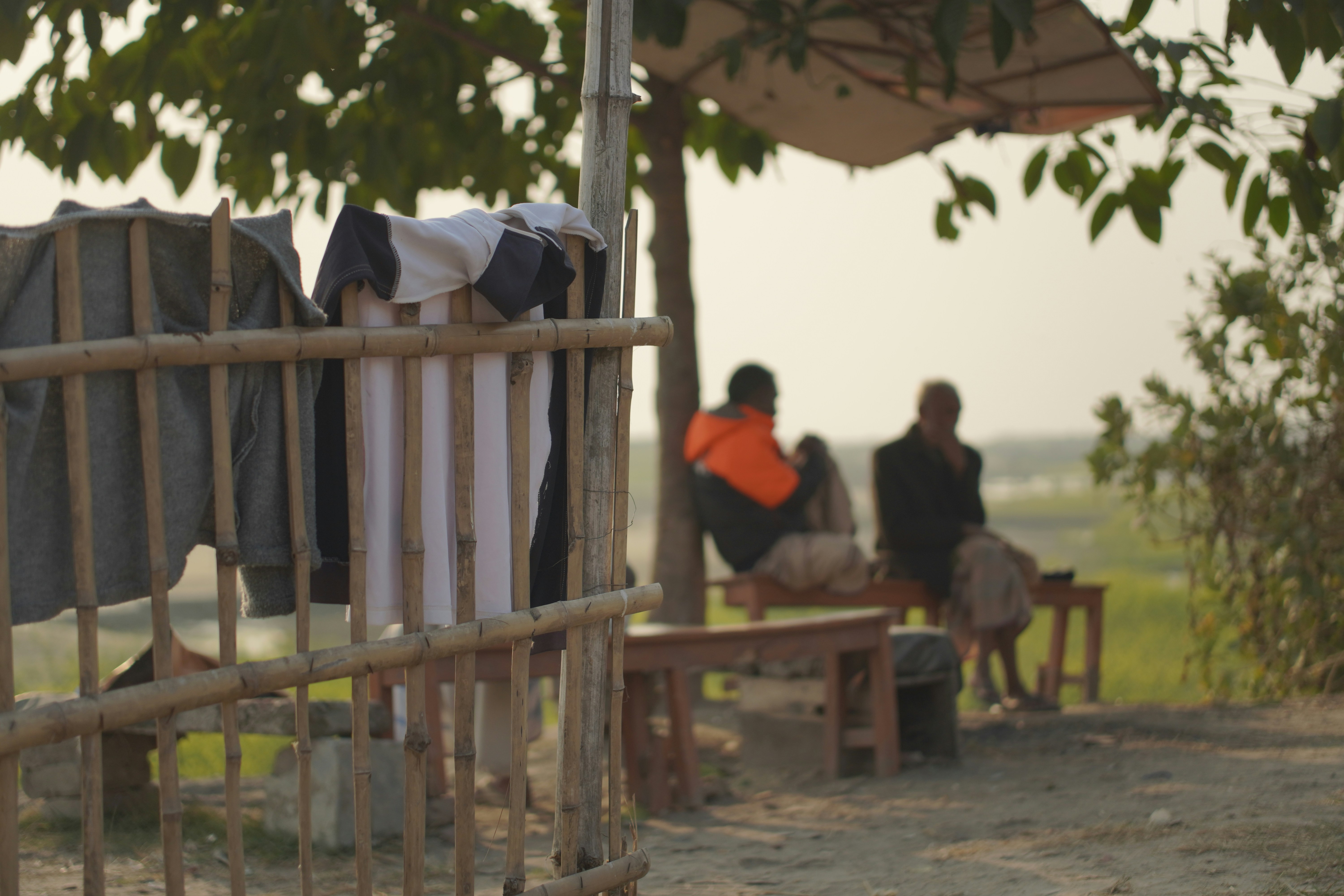 People drinking water and trying to stay cool under a makeshift shade structure in a hot, dusty rural area.
