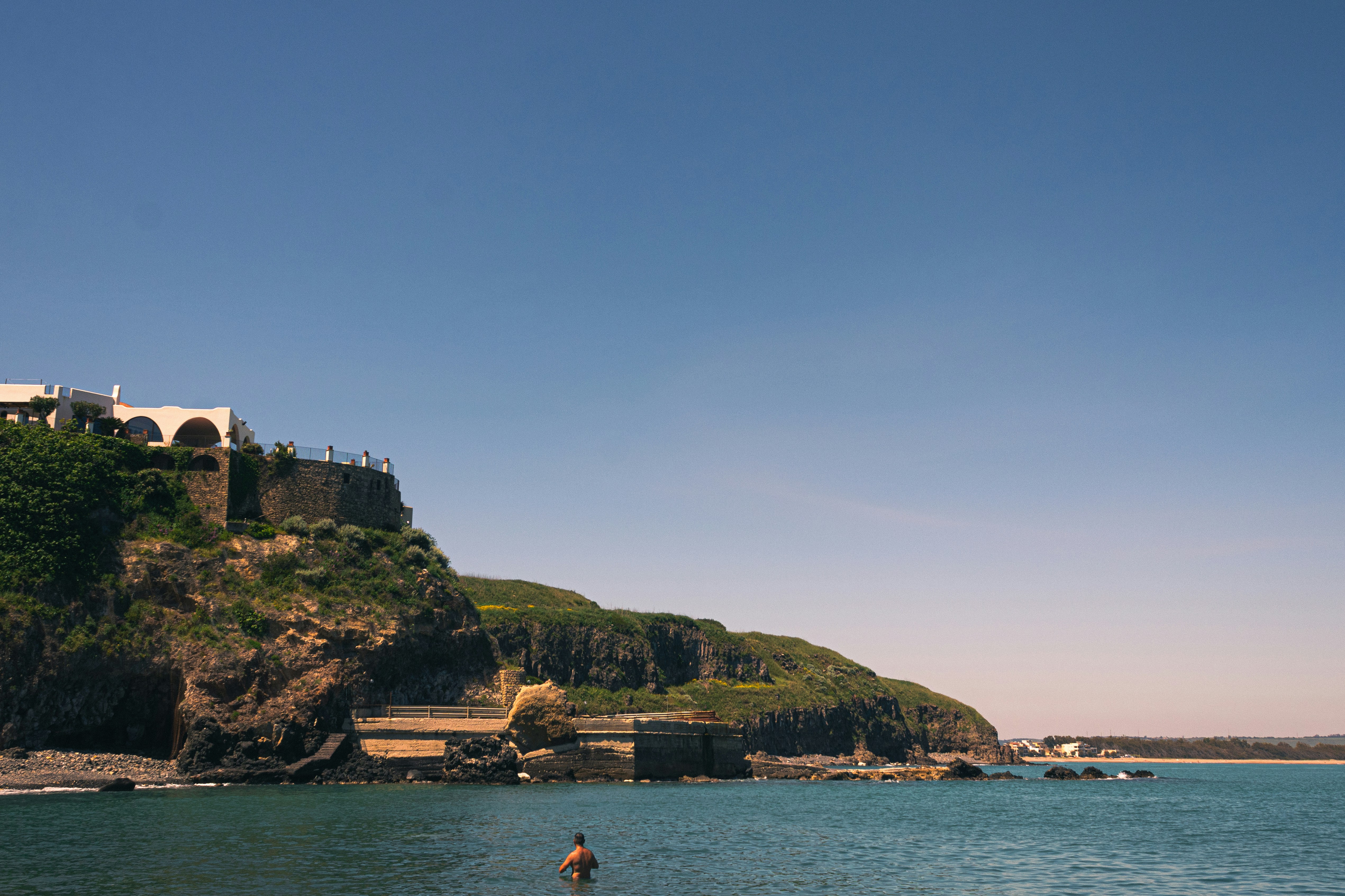 A person stands in the ocean near a cliffside building