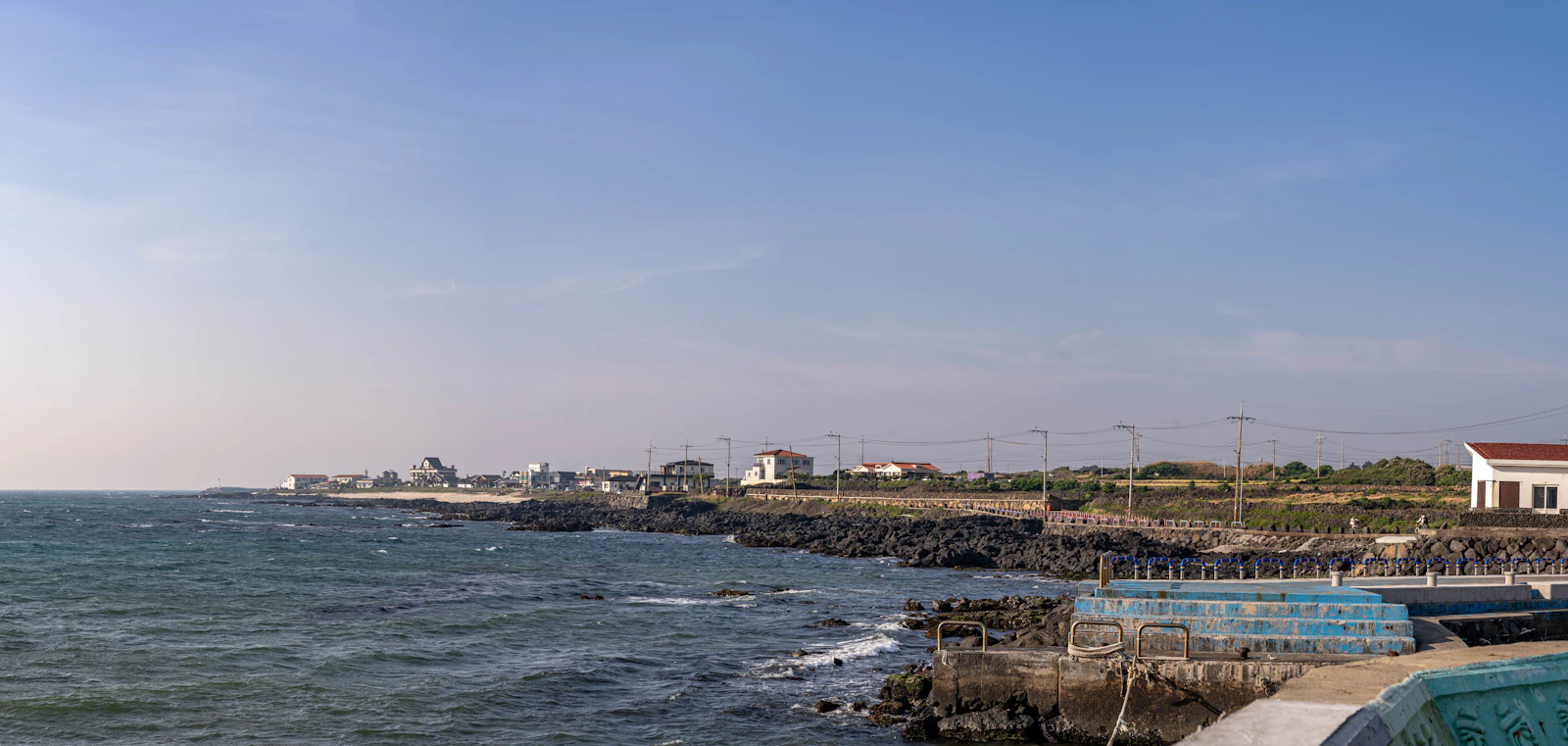 Rocky Jeju shoreline with low buildings beneath a bright sky