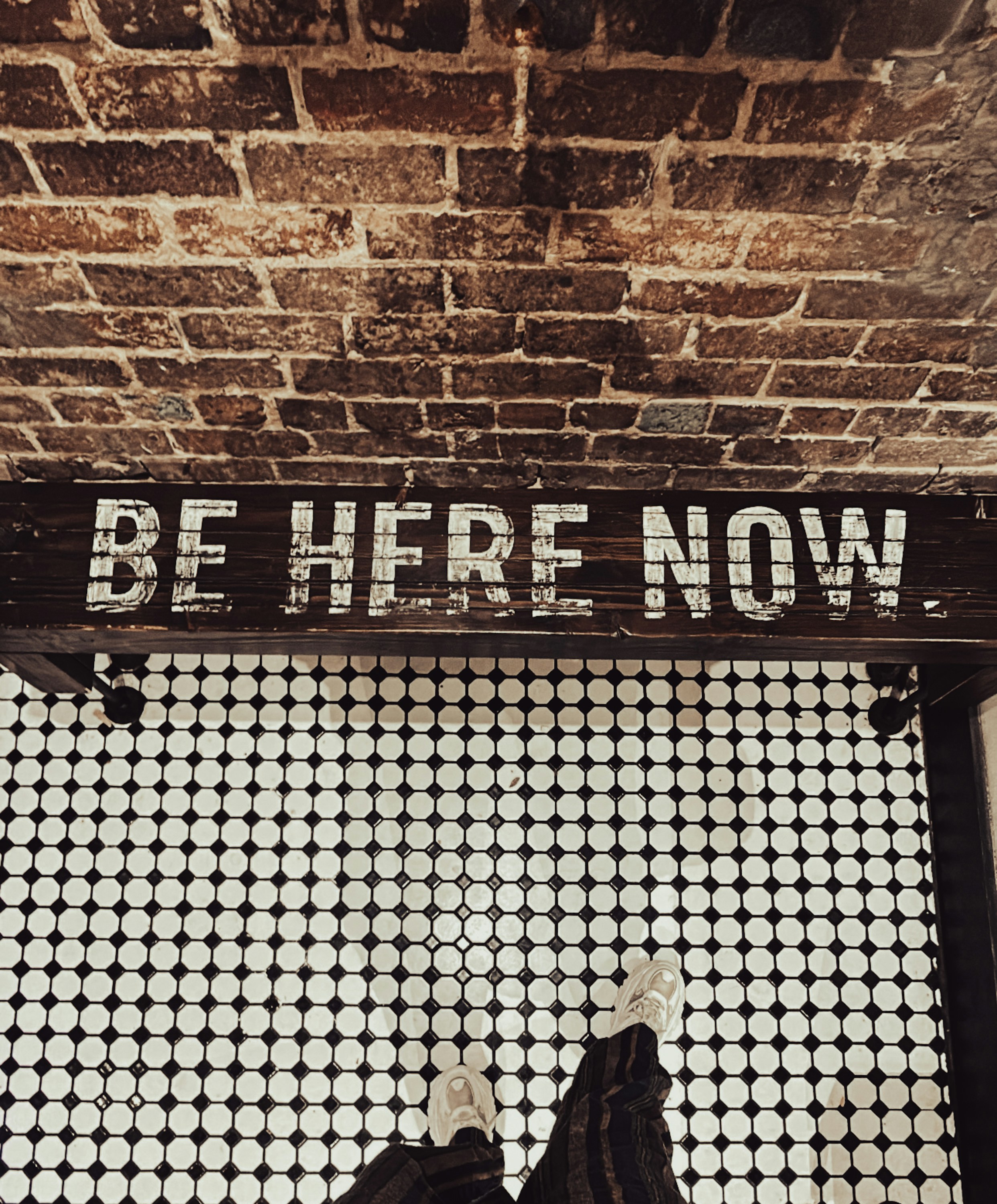 Sign says be here now above tiled floor