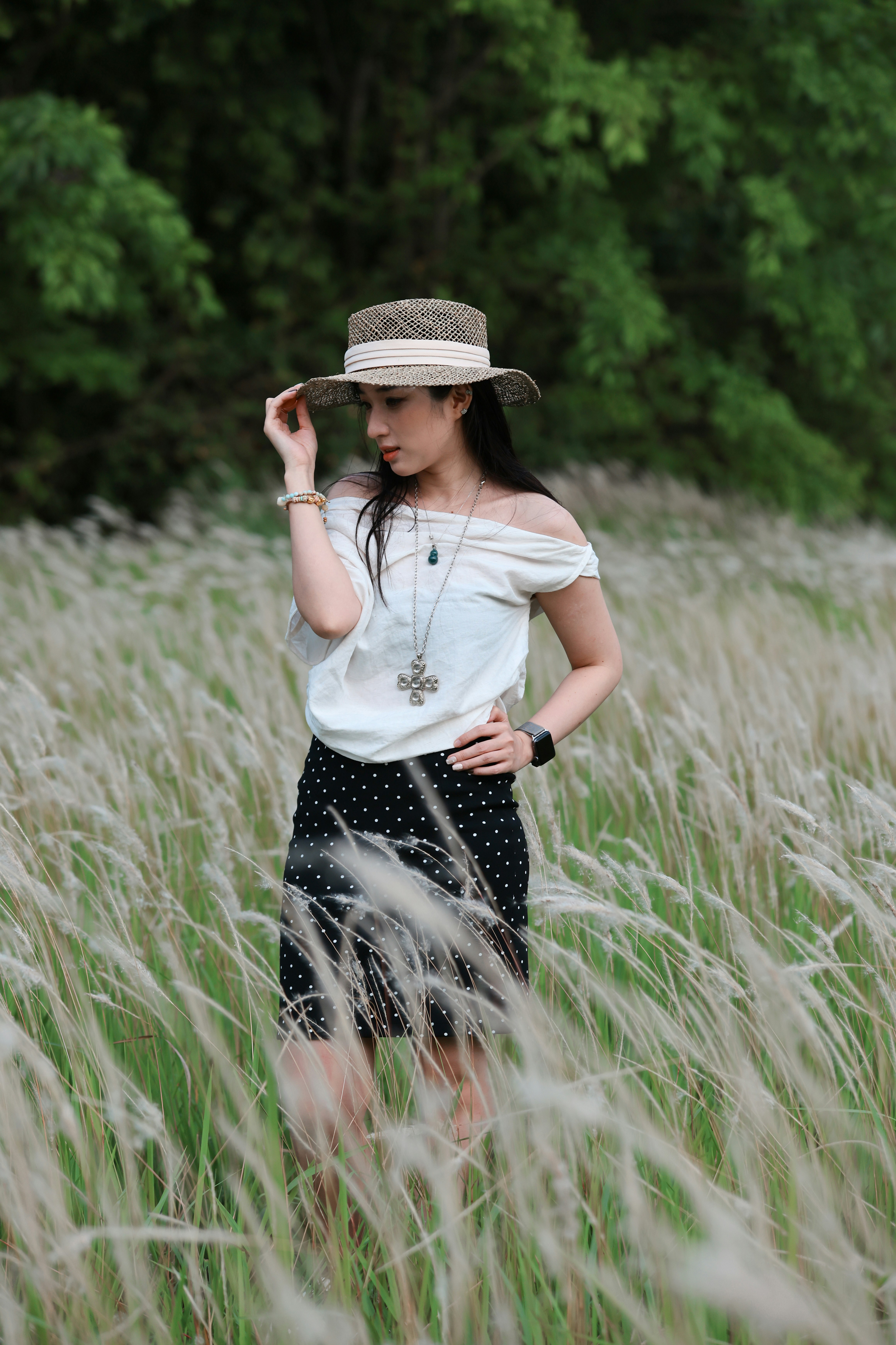 A woman in a white off‑the‑shoulder top and polka-dot skirt stands in a tall grass field, wearing a straw hat and layered jewelry; she gazes downward while one hand lifts the hat, creating a contemplative, summer mood.