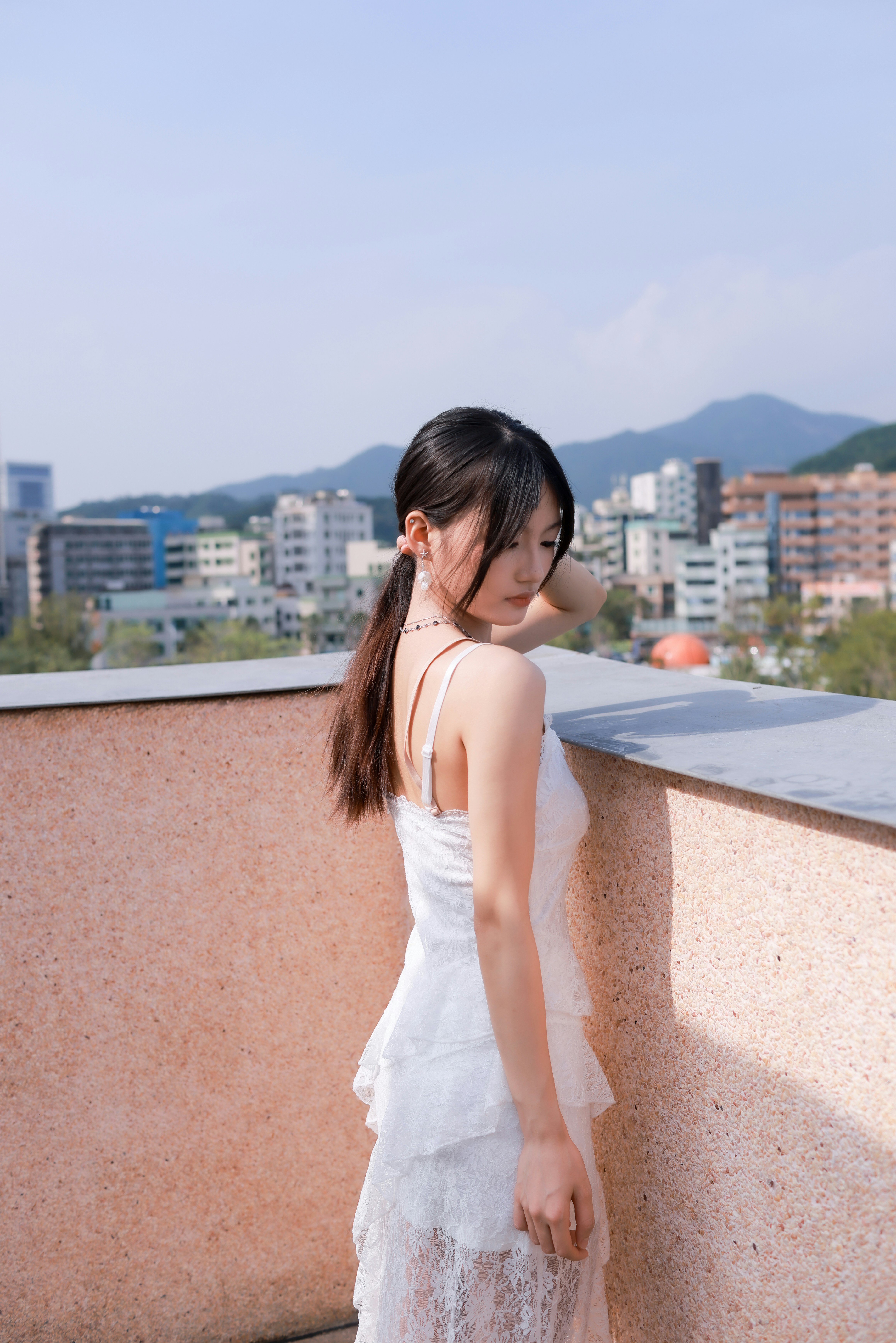 A young woman leans against a rooftop ledge in a white lace dress, with a distant city and hills in the background.