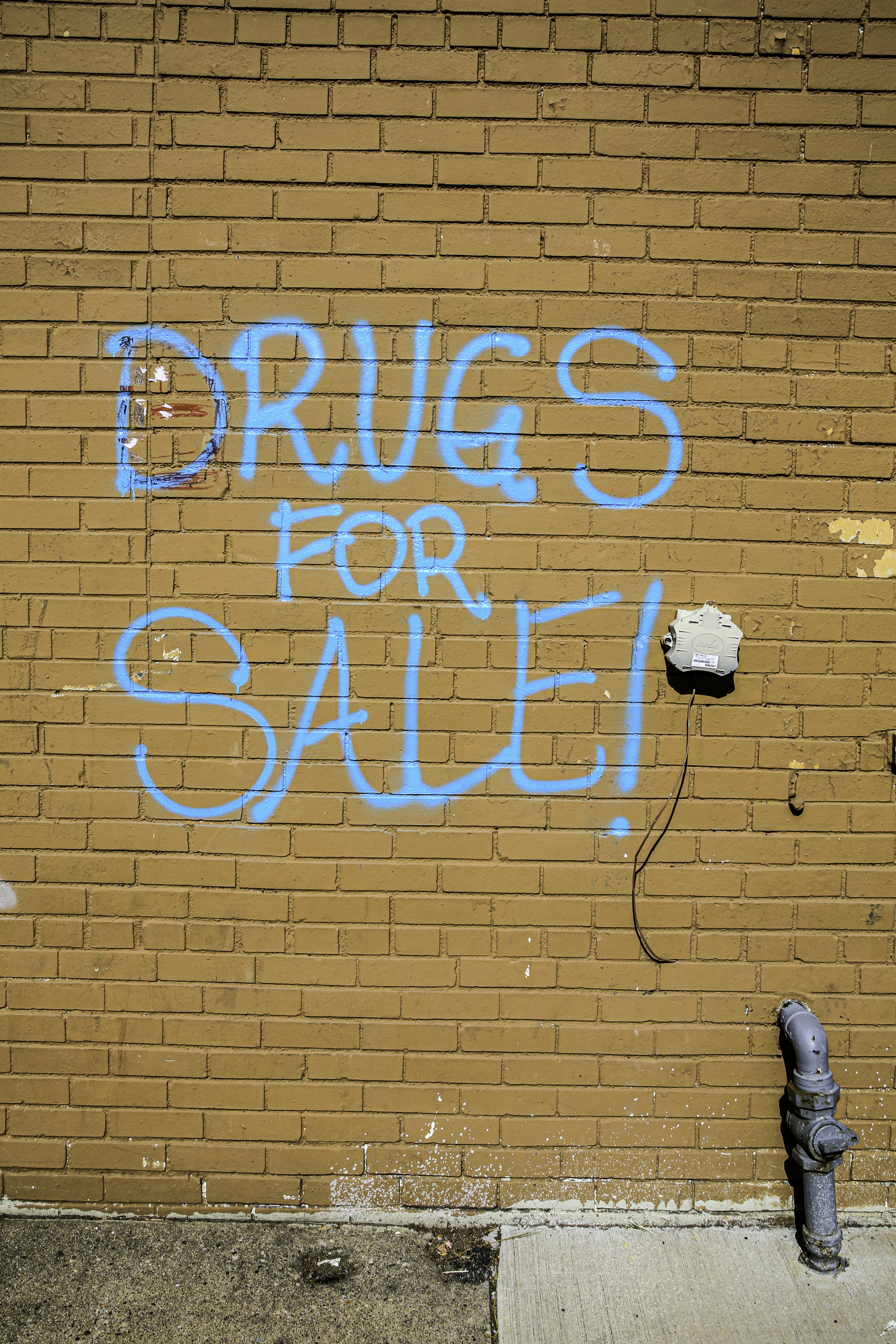 Blue graffiti 'drugs for sale!' on a brick wall.